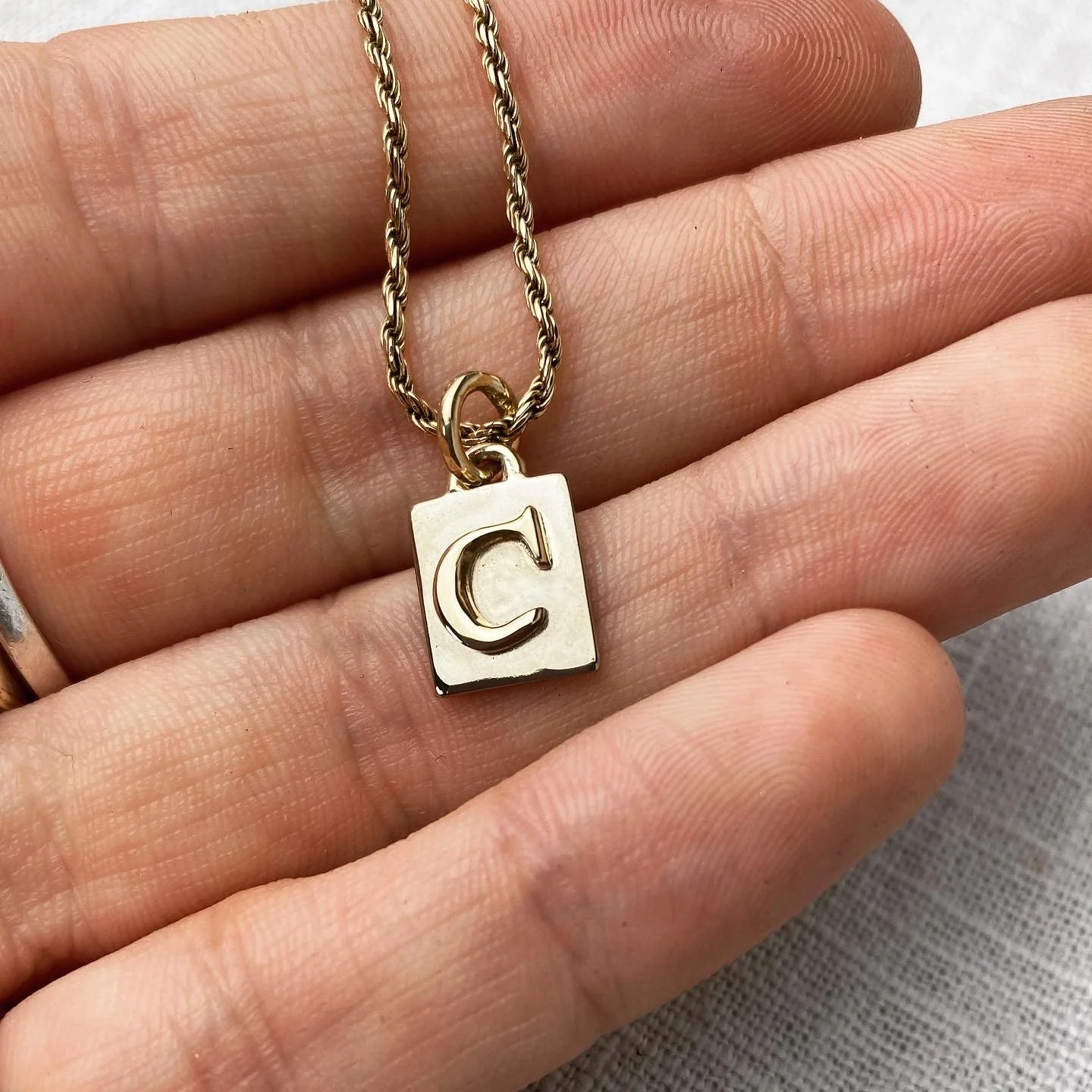 Close-up of a gold necklace with a pendant featuring a letter 'C' engraved on a square background, held against a person's hand.