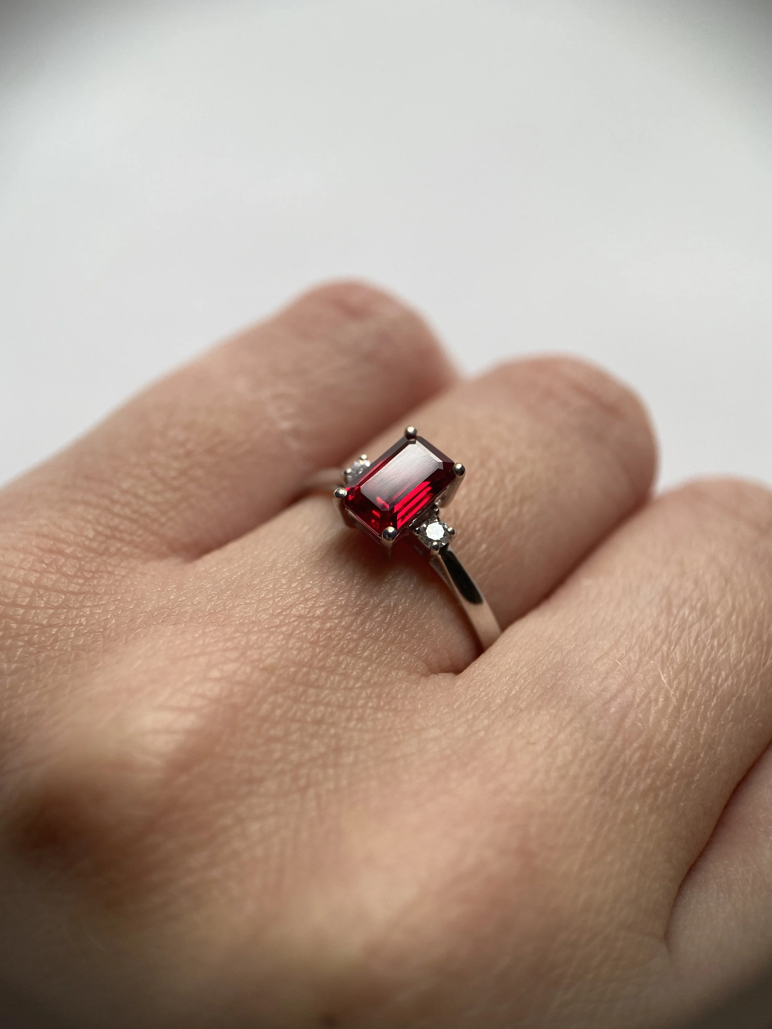 Close-up of a hand wearing a silver ring with a large rectangular red gemstone and small clear stones on each side, against a plain white background.