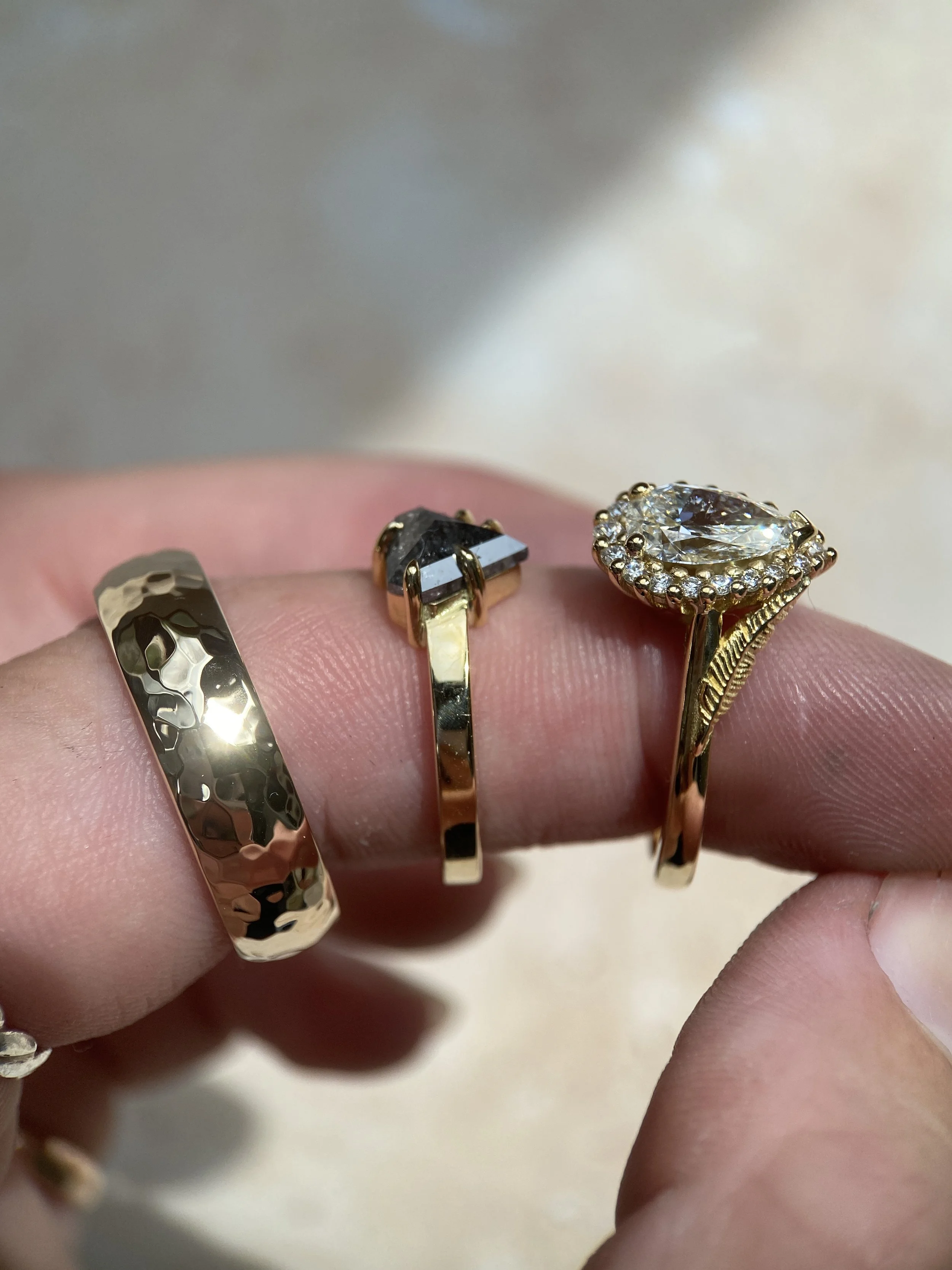 Close-up of a hand wearing three rings: a hammered gold band, a gold ring with a dark gemstone, and a gold ring with a large clear gemstone surrounded by smaller stones.