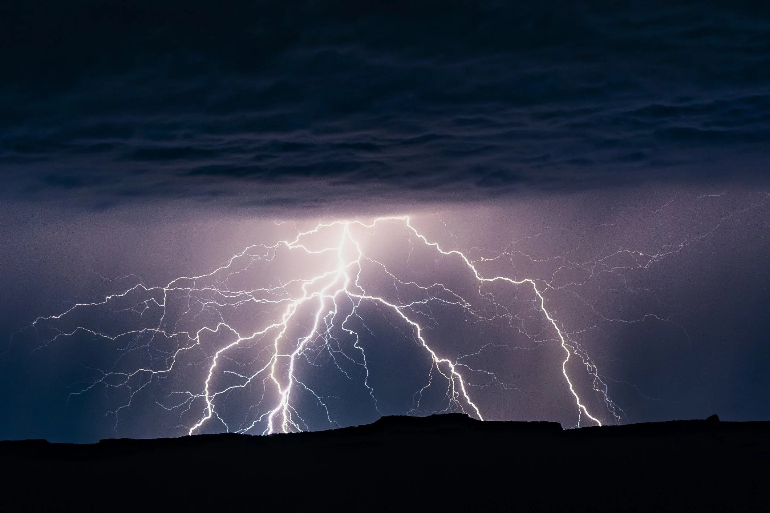 A massive branching bolt strikes behind the silhouetted Vermillion Cliffs.