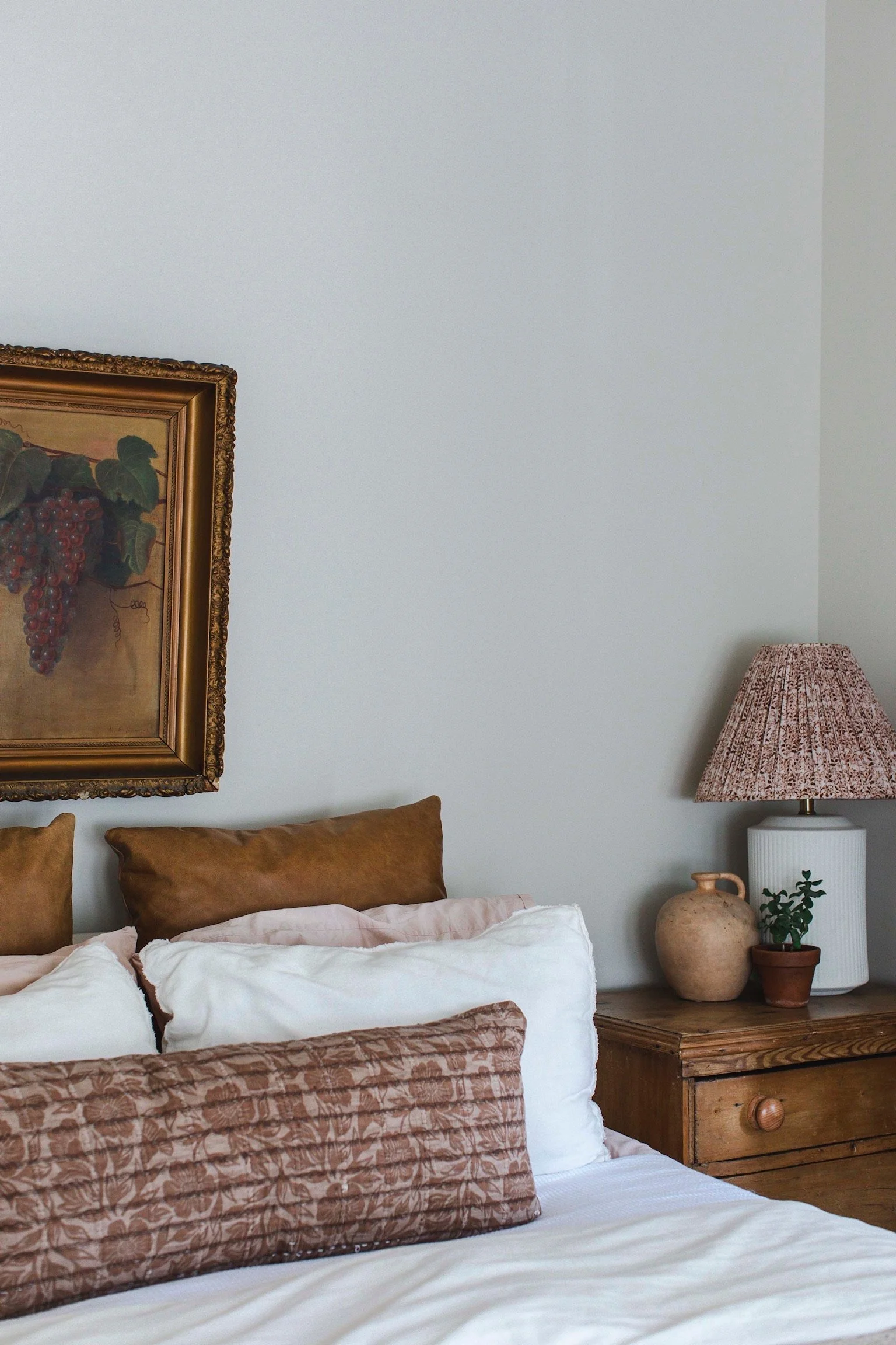 A cozy bedroom corner with a bed, pillows, a wooden nightstand, a table lamp, a small potted plant, a decorative clay vase, and a framed painting of grapes on the wall.