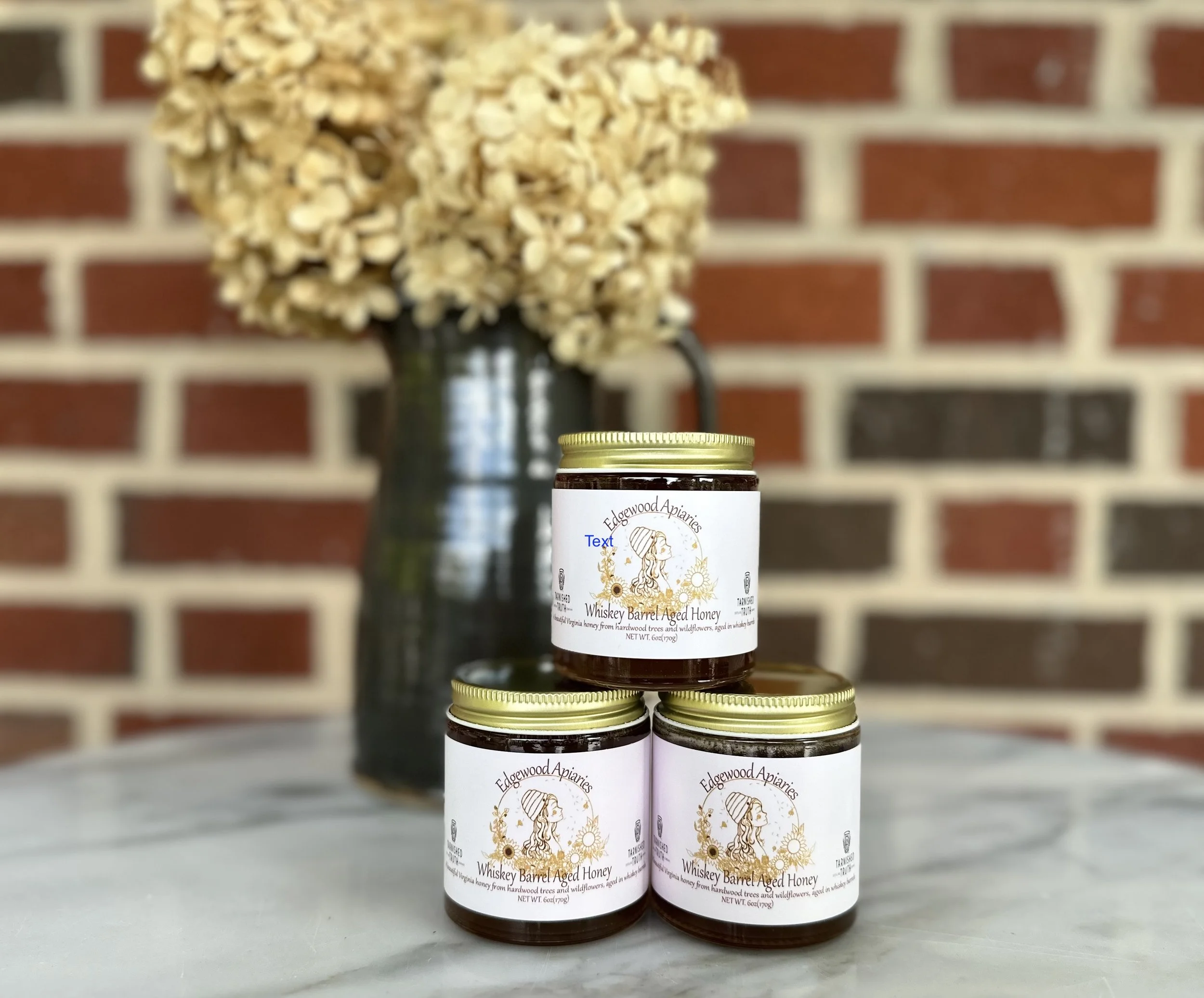 A young woman with brown hair, glasses, and a blue T-shirt standing against a red brick wall, holding a bar of handmade soap.