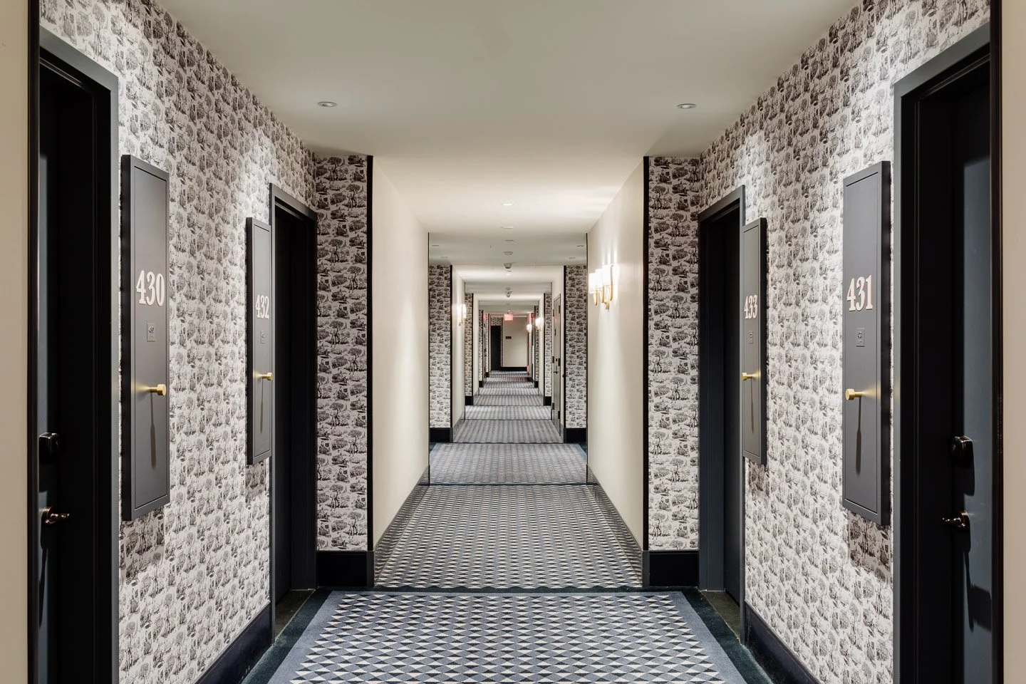 A hotel hallway with patterned carpet, brick-patterned wallpaper on the sides, and black doors with white numbers on the left side.