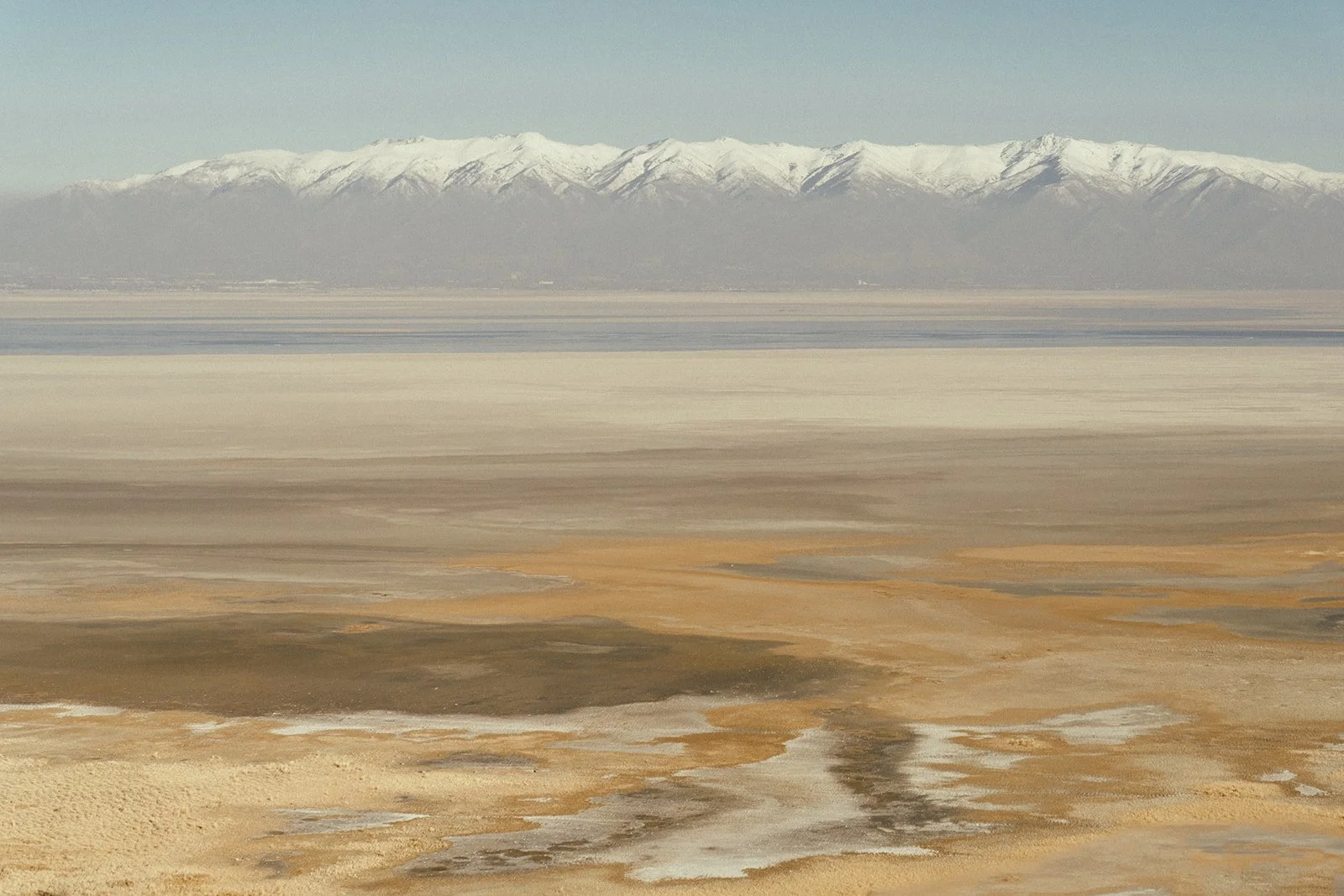 Scenic view of snow-capped mountains in the distance, with a flat, colorful salt flat and dried mud in the foreground.