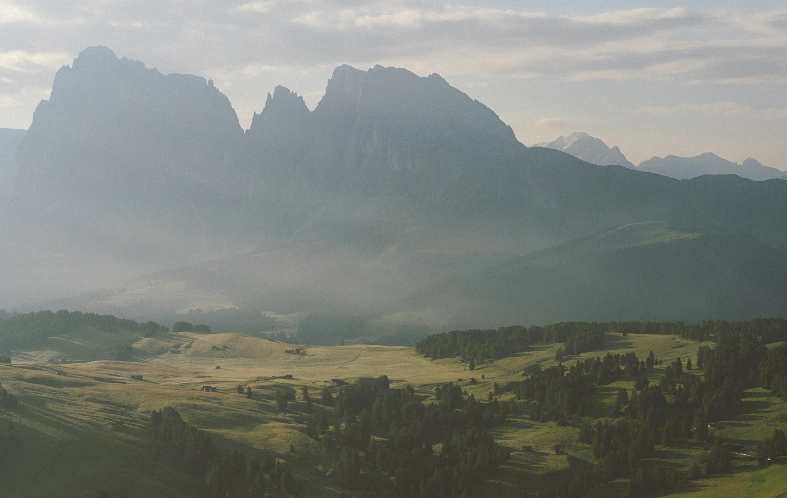 Scenic landscape of rolling green hills with trees and roads, and a mountain range in the background under a cloudy sky.