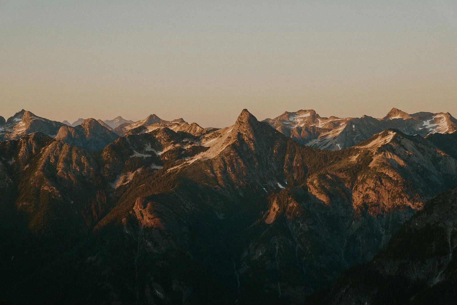 A mountain range at sunset with rugged peaks and patches of snow, under a clear sky.