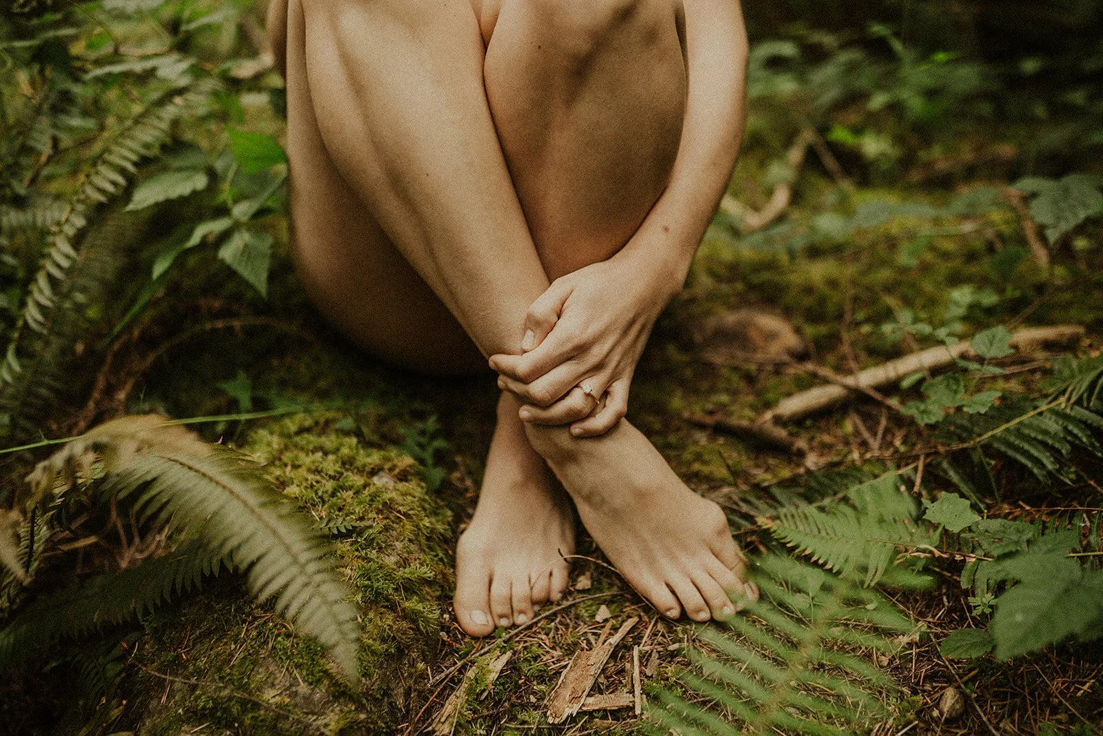 A person sitting on the forest floor with their knees drawn up, holding their legs with both hands. The person is barefoot and surrounded by green ferns and moss.