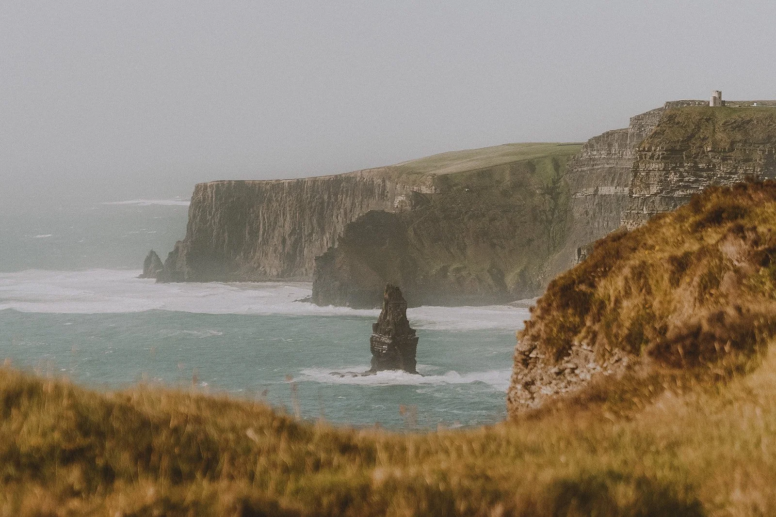 Cliffs along a rugged coastline with a tall sea stack in the foreground and grassy foreground, misty atmosphere, and a small building near the top of the cliffs.