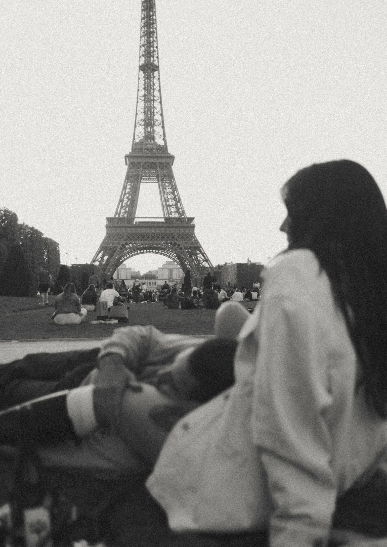 A black and white photo of the Eiffel Tower in Paris with a woman sitting in the foreground, overlooking the scene, along with other people relaxing on the grass.