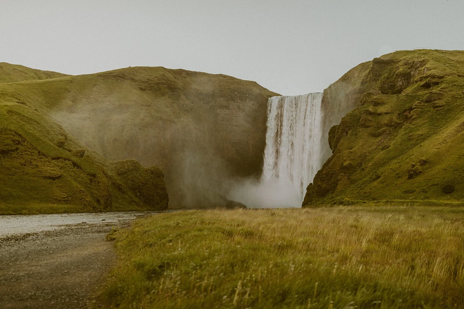 A waterfall cascading down a cliff surrounded by grassy hills on a cloudy day.
