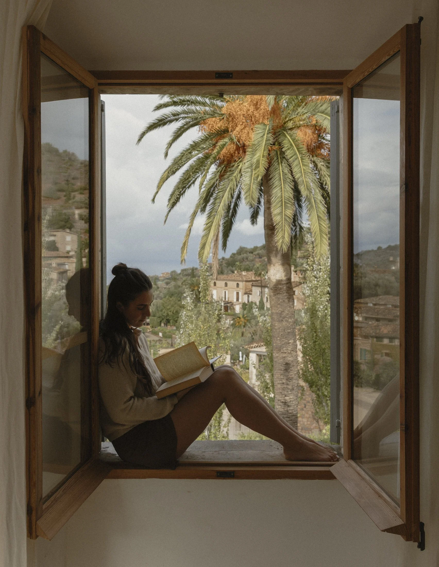 A woman sitting on a windowsill reading a book with a view of a palm tree and hillside houses outside.