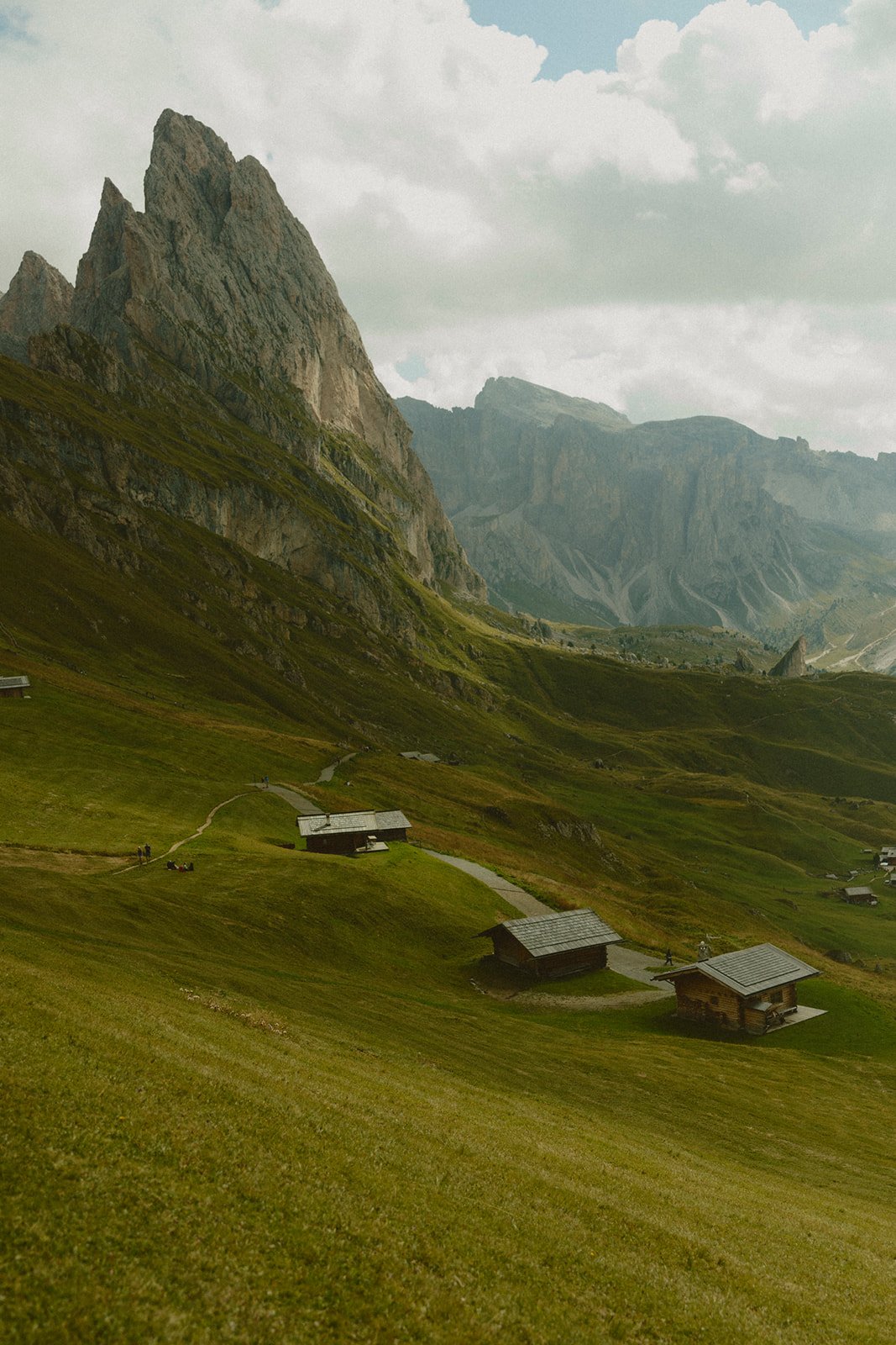 Landscape with green rolling hills, scattered wooden cabins, and towering mountain peaks in the background under a cloudy sky.