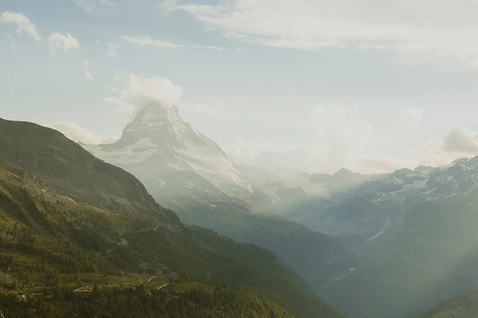 Scenic view of snow-capped mountain with clouds, green rolling hills, and forested valleys.
