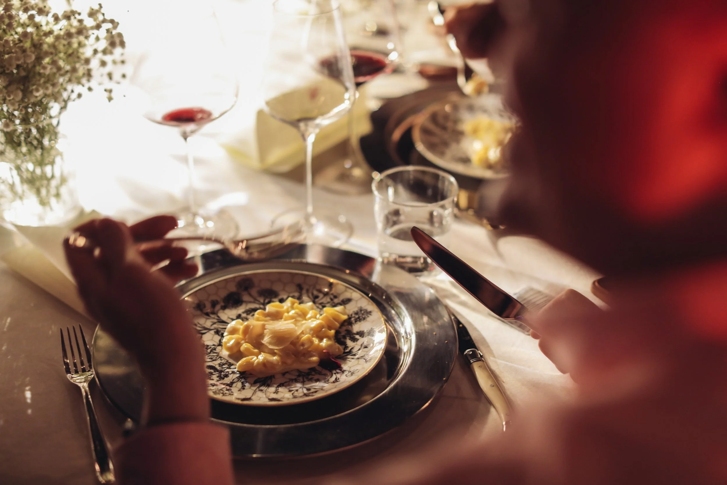 People are dining at a formal dinner table, with wine glasses and a plate of pasta in the foreground. There is a centerpiece with flowers and a cheese platter in the background.