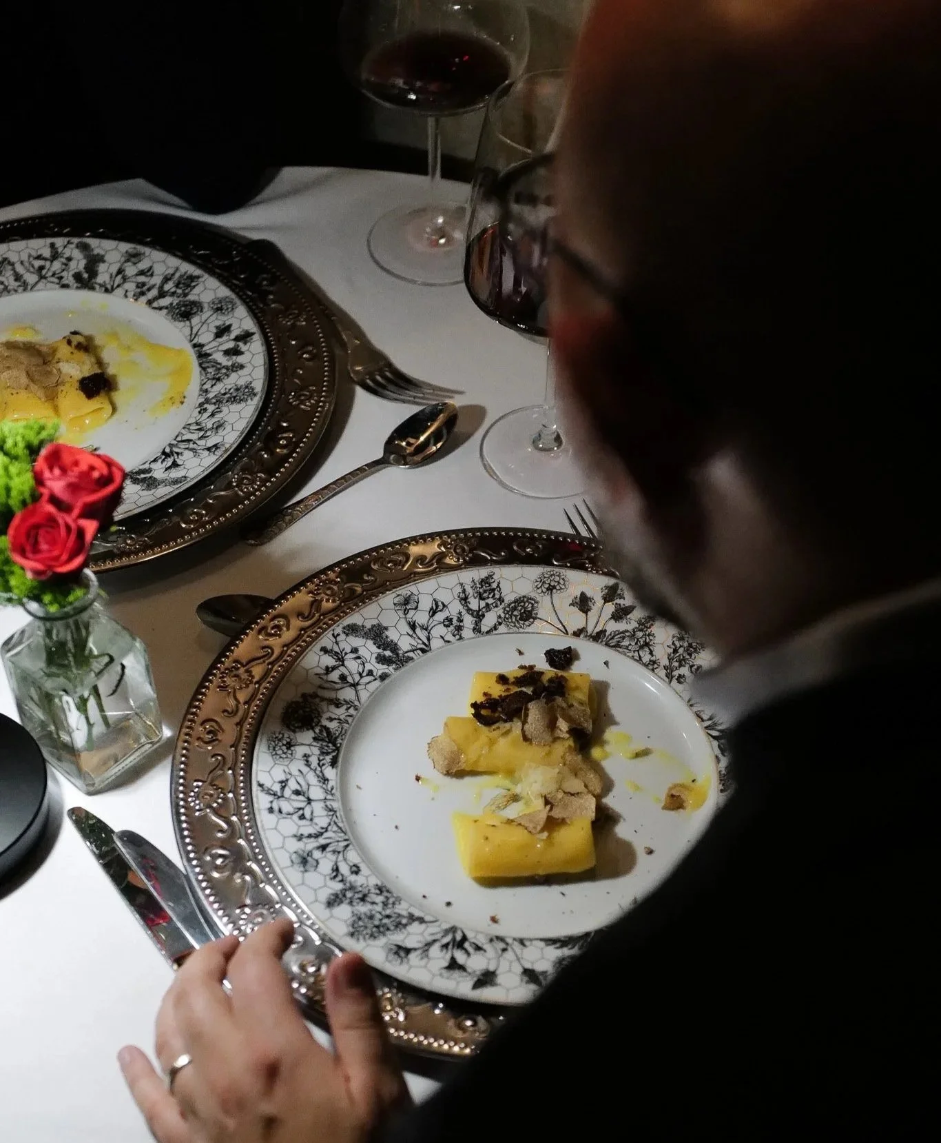 A person at a formal dinner table with ornate plates, a glass of red wine, and a small bouquet of red roses in a glass vase. The person is holding a serving tray with a plated dish of pasta topped with shaved truffles.