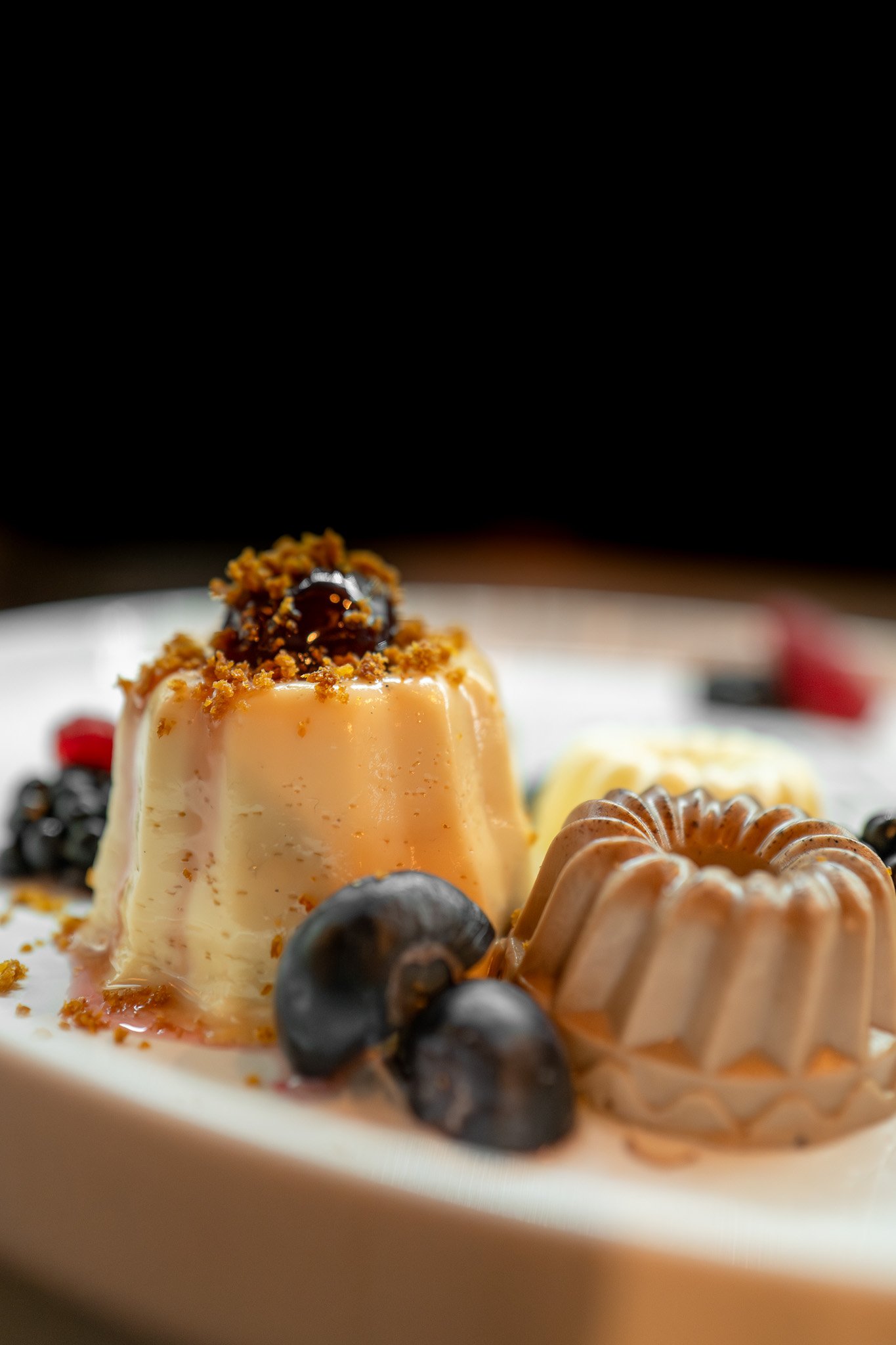 Close-up of a caramel custard dessert topped with crushed cookies and a cherry, surrounded by blueberries and other desserts on a white plate.
