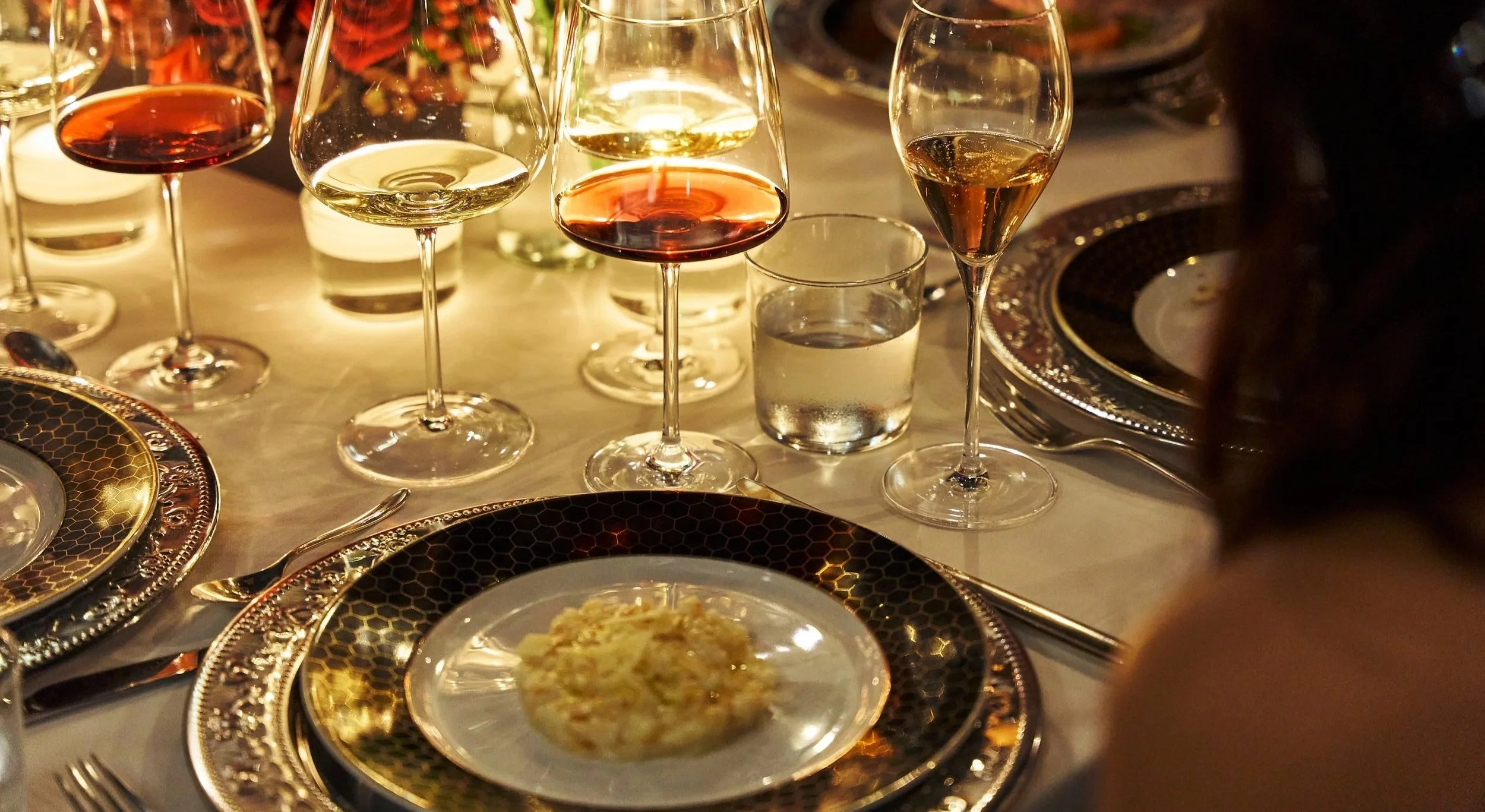 An elegant dinner table setting with multiple wine glasses filled with red and white wine, ornate gold and black patterned plates, a small plate of food in the foreground, and silverware, all under warm ambient lighting.