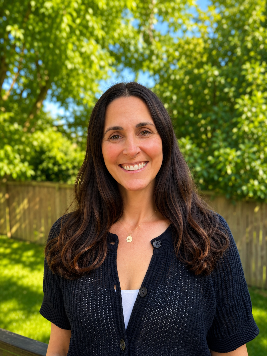 Smiling woman with long dark hair wearing a black knitted cardigan and a necklace, standing outdoors in a backyard with green trees and a wooden fence in the background.