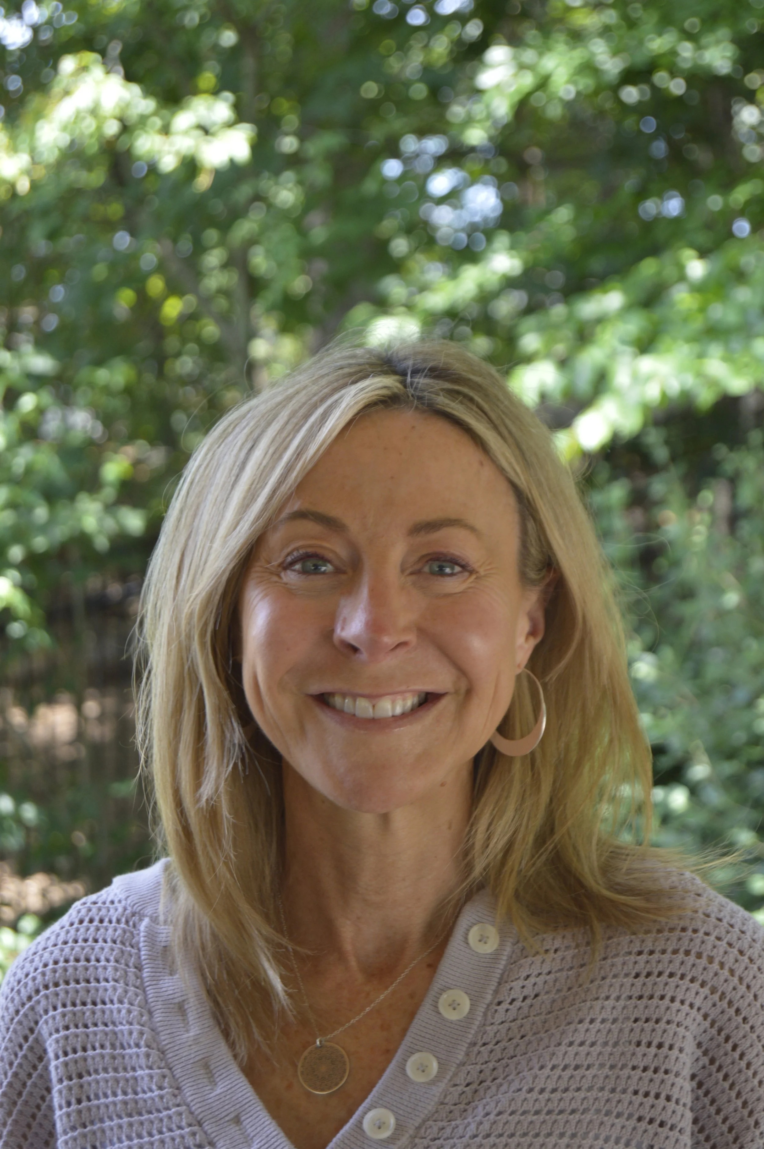 Smiling woman with blonde hair and hoop earrings outdoors, wearing a light-colored knit top with buttons, in front of a leafy green background.