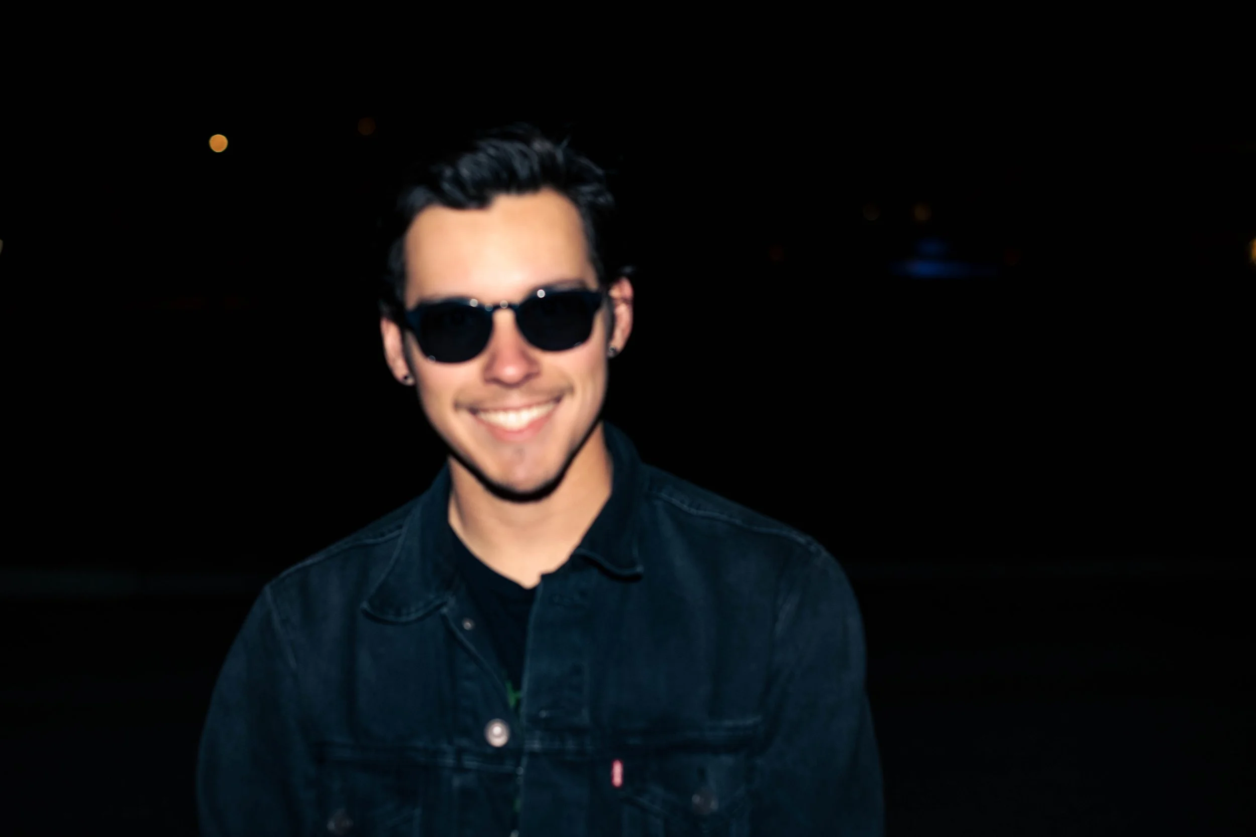 Young man smiling at night, wearing sunglasses and a dark denim jacket.