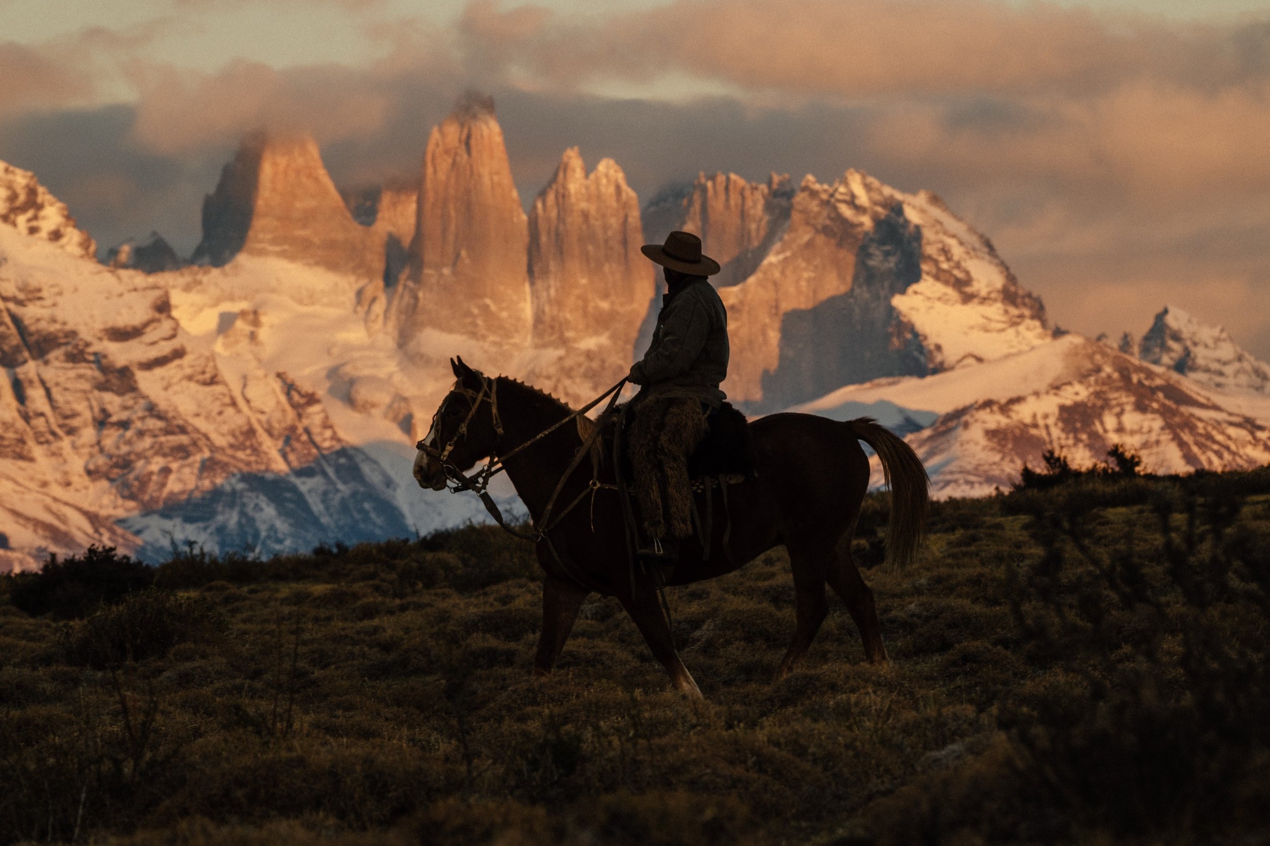 Person riding a horse through a mountainous landscape at sunset, with snow-capped peaks in the background.