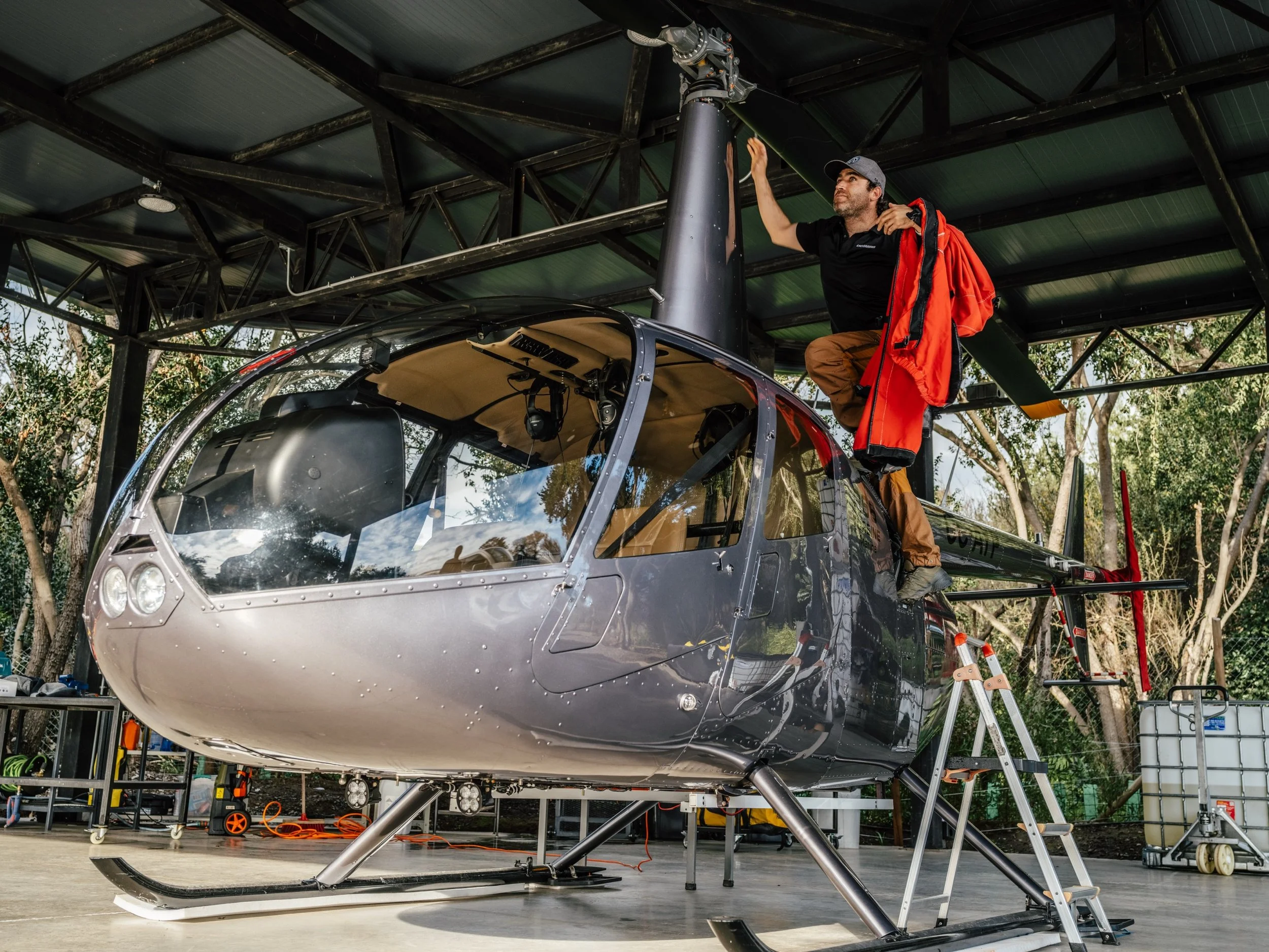 A person standing on top of a black helicopter, working on the rotor mechanism under a metal shelter, surrounded by trees and equipment.