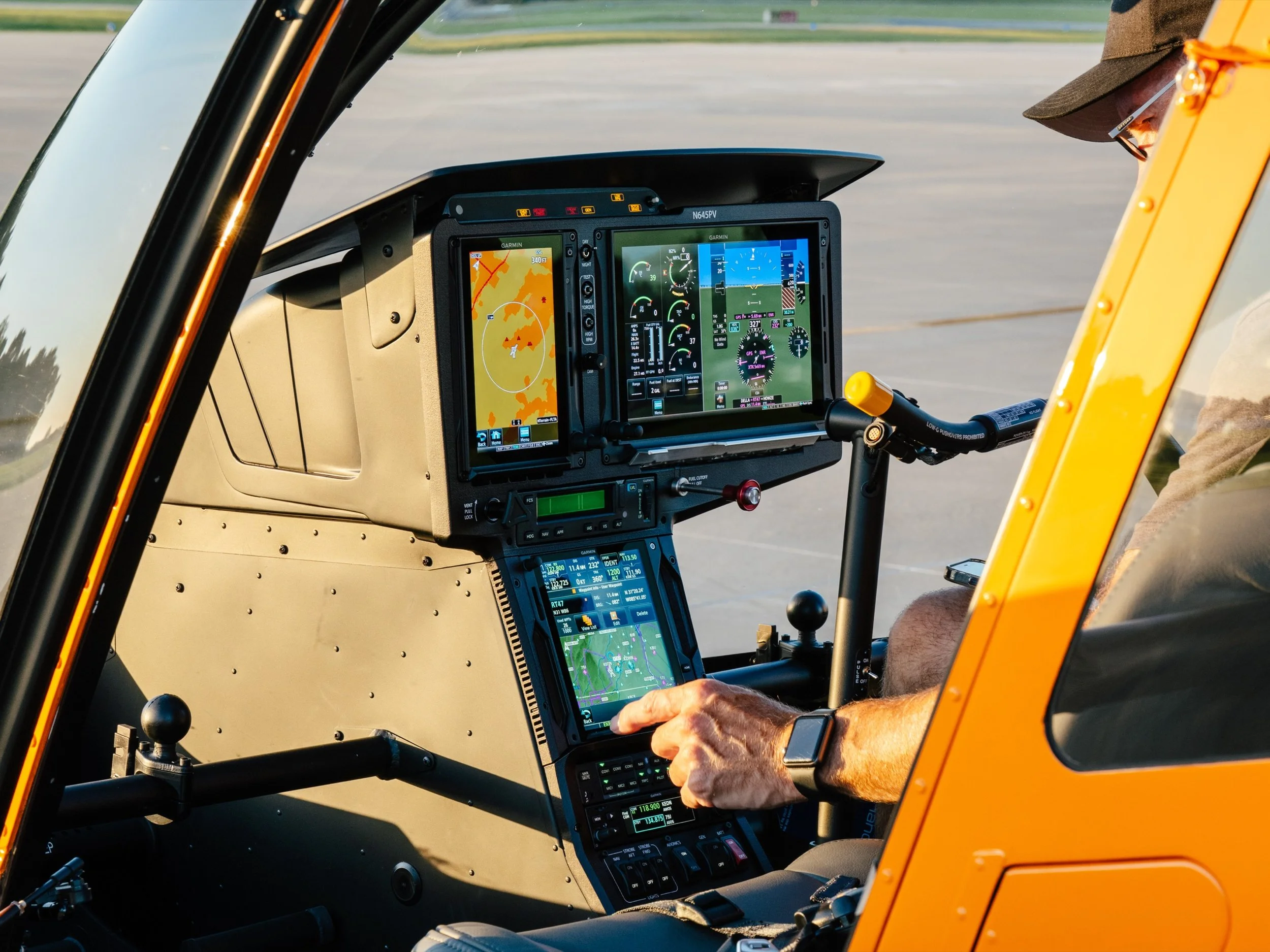 Close-up of a pilot's hands operating a glass cockpit helicopter with multiple digital screens displaying navigation, altitude, and weather information, parked on the tarmac.