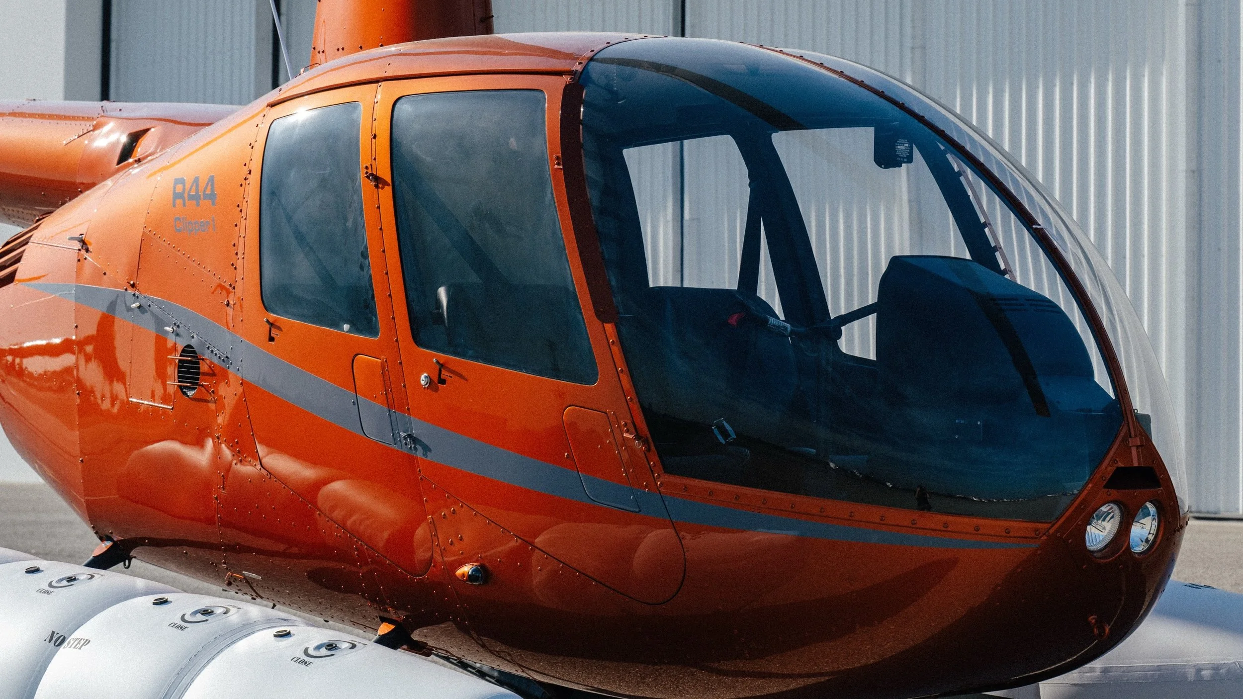 A close-up of an orange helicopter with the label 'R44 Clipper' on the side, parked outside a hangar.