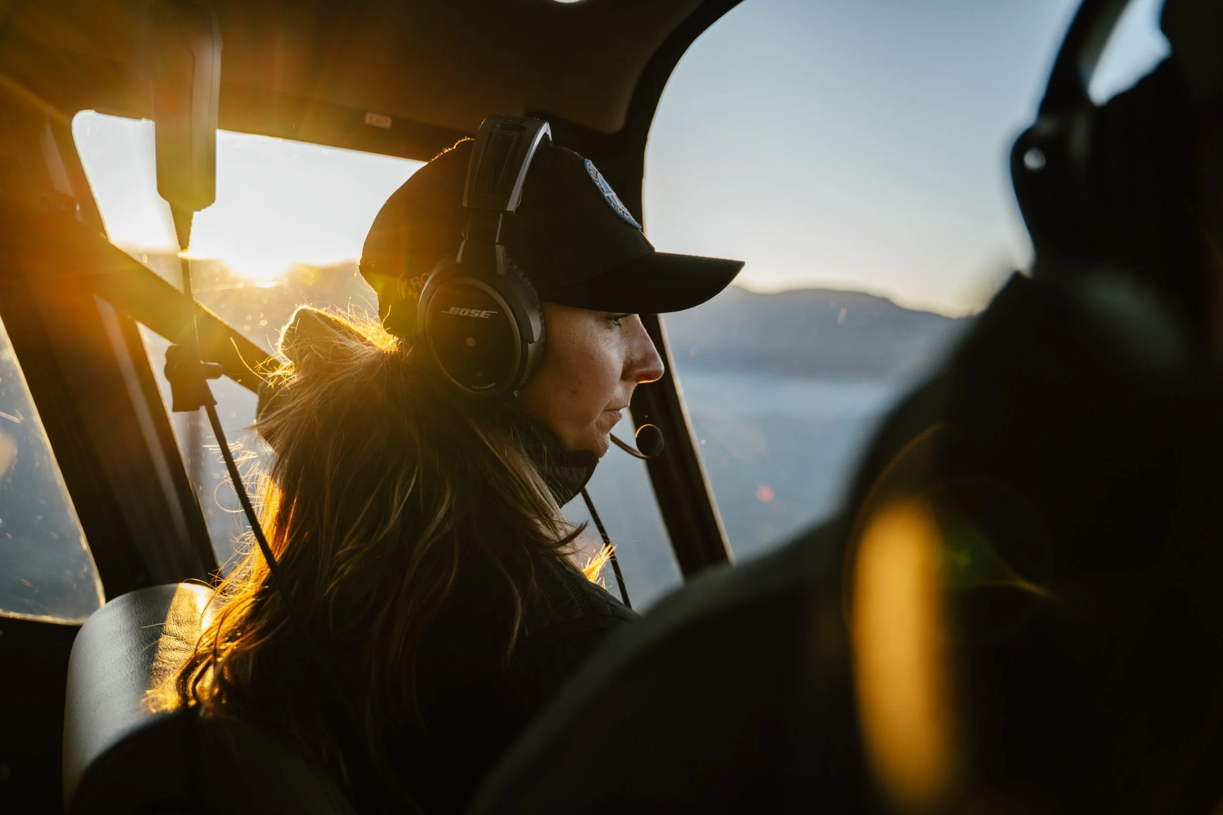 A woman wearing a headset and a baseball cap, sitting inside a cockpit with sunlight shining through the window, with mountains outside in the background.