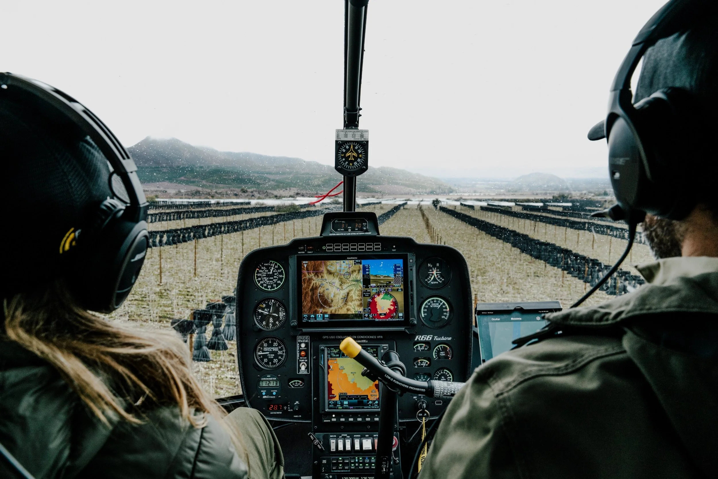 View from inside a helicopter cockpit showing two pilots, with various instruments and GPS screens, flying over a vineyard with mountains in the background.