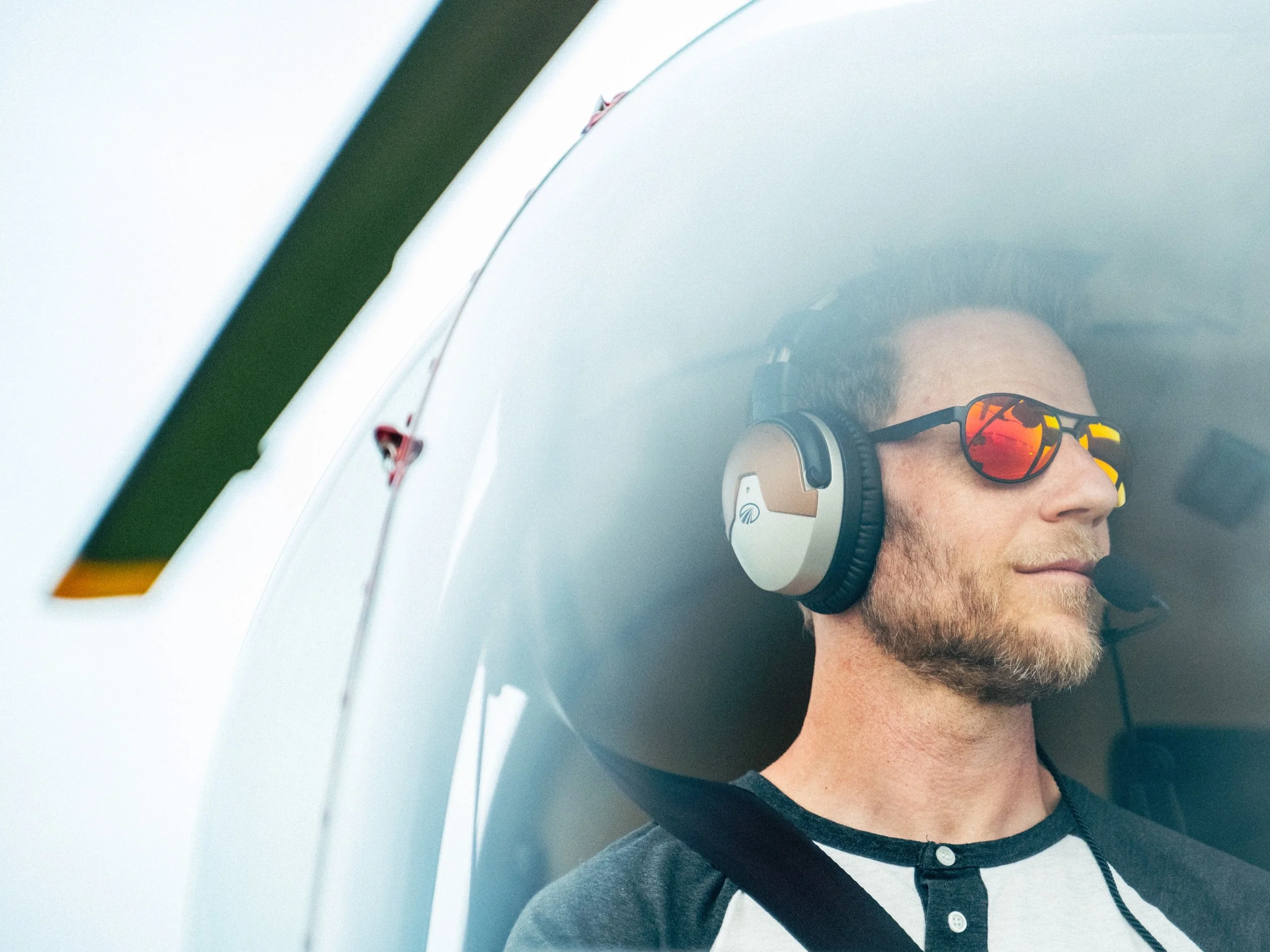 A man with a beard wearing sunglasses and headphones, lying against the side of a white aircraft with a blurred background.