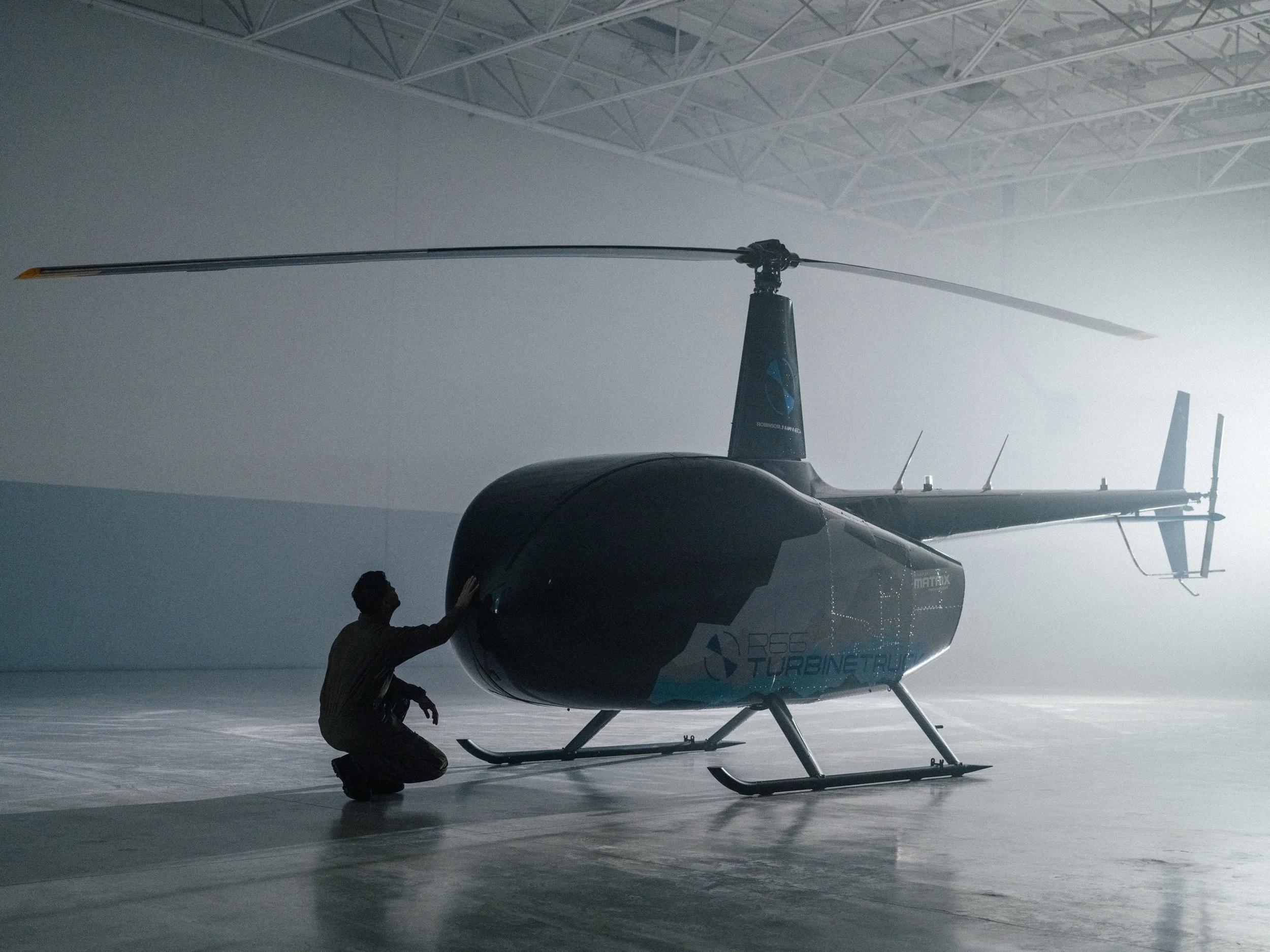 A person kneeling next to a small helicopter inside an industrial hangar with high ceilings and exposed trusses. The helicopter has a sleek black and white body with a prominent rotor on top and the words "Turbine" visible on the side.