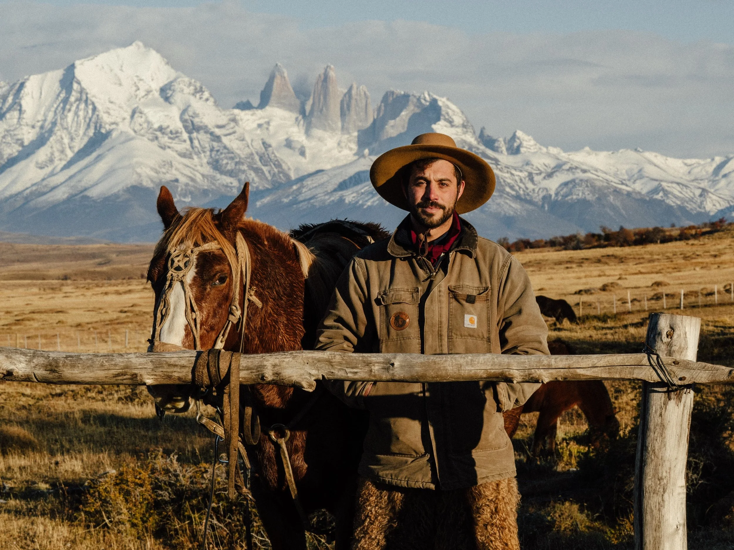 A man with a beard wearing a tan jacket and large wide-brimmed hat standing behind a wooden fence with a brown and white horse, set against a backdrop of snow-capped mountains and cloudy sky.