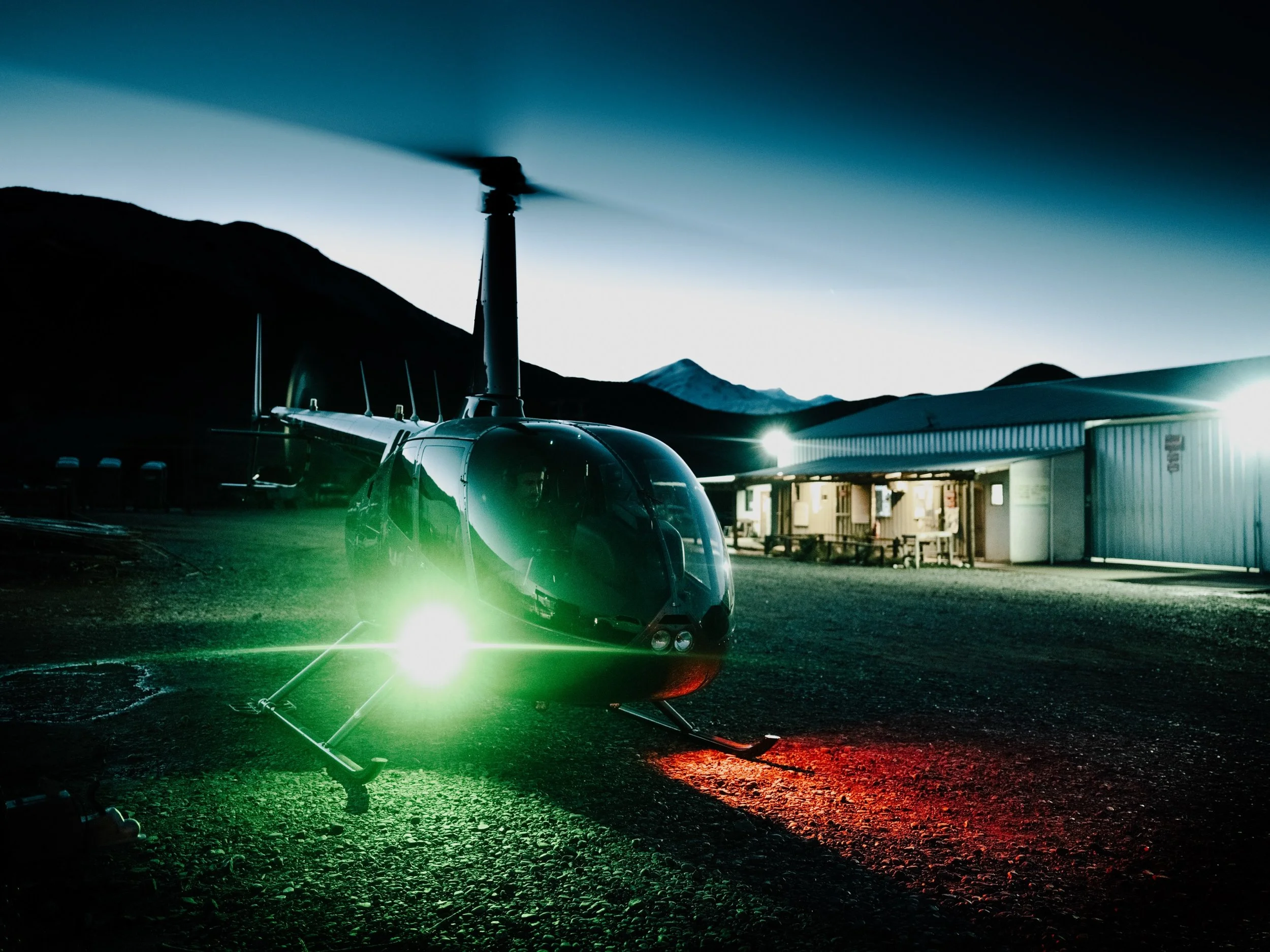 A helicopter with bright lights on the ground at dusk, mountains in the background, and a building nearby.