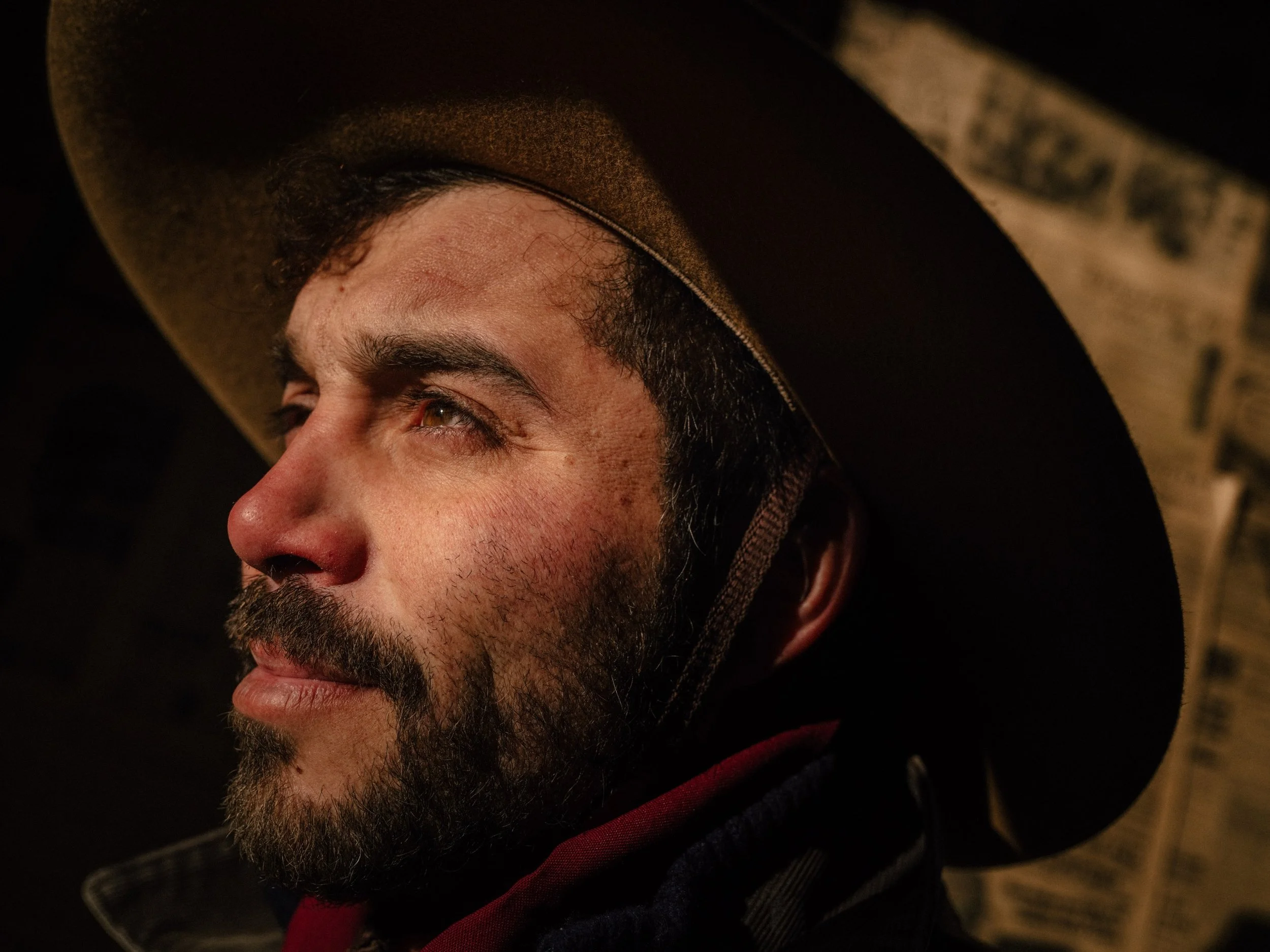 Close-up of a man's face, showing his eyes, eyebrows, nose, and beard, wearing a brown hat. The background is dark.