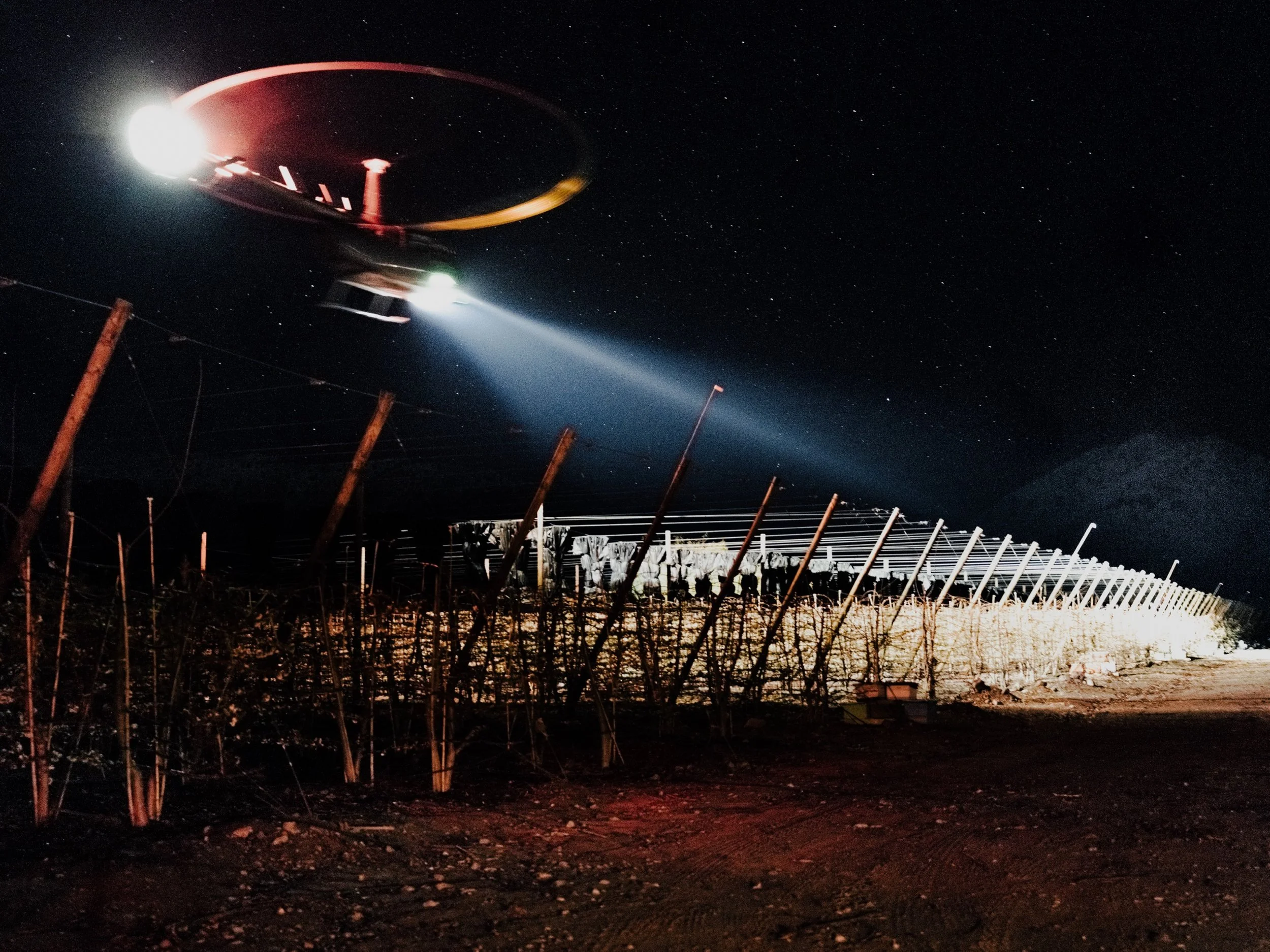 A helicopter flying at night with bright lights and a searchlight shining downward. The scene includes starry sky, power lines, and damaged fence or barrier in the foreground.