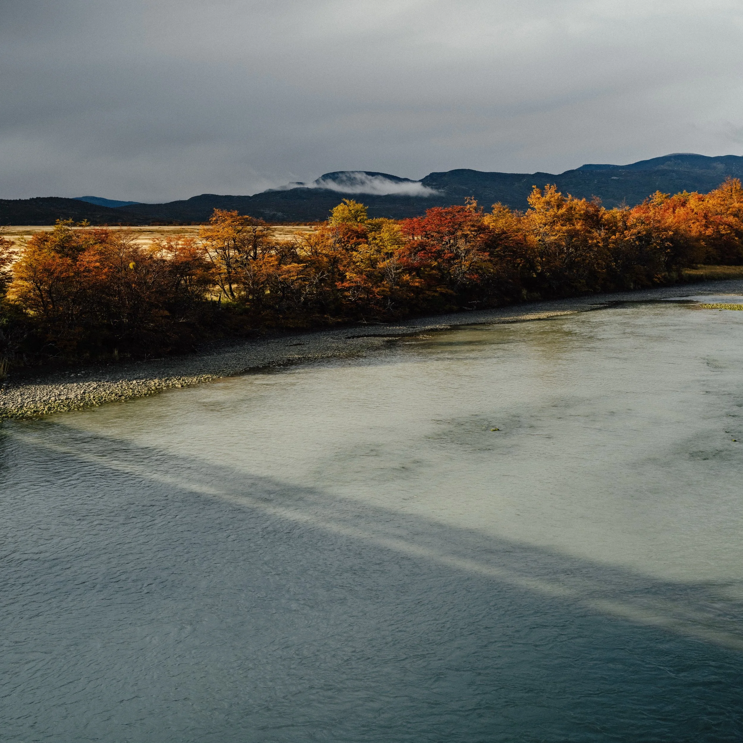 A landscape scene with a river in the foreground, surrounded by trees with autumn foliage, mountains in the background, and a cloudy sky above.