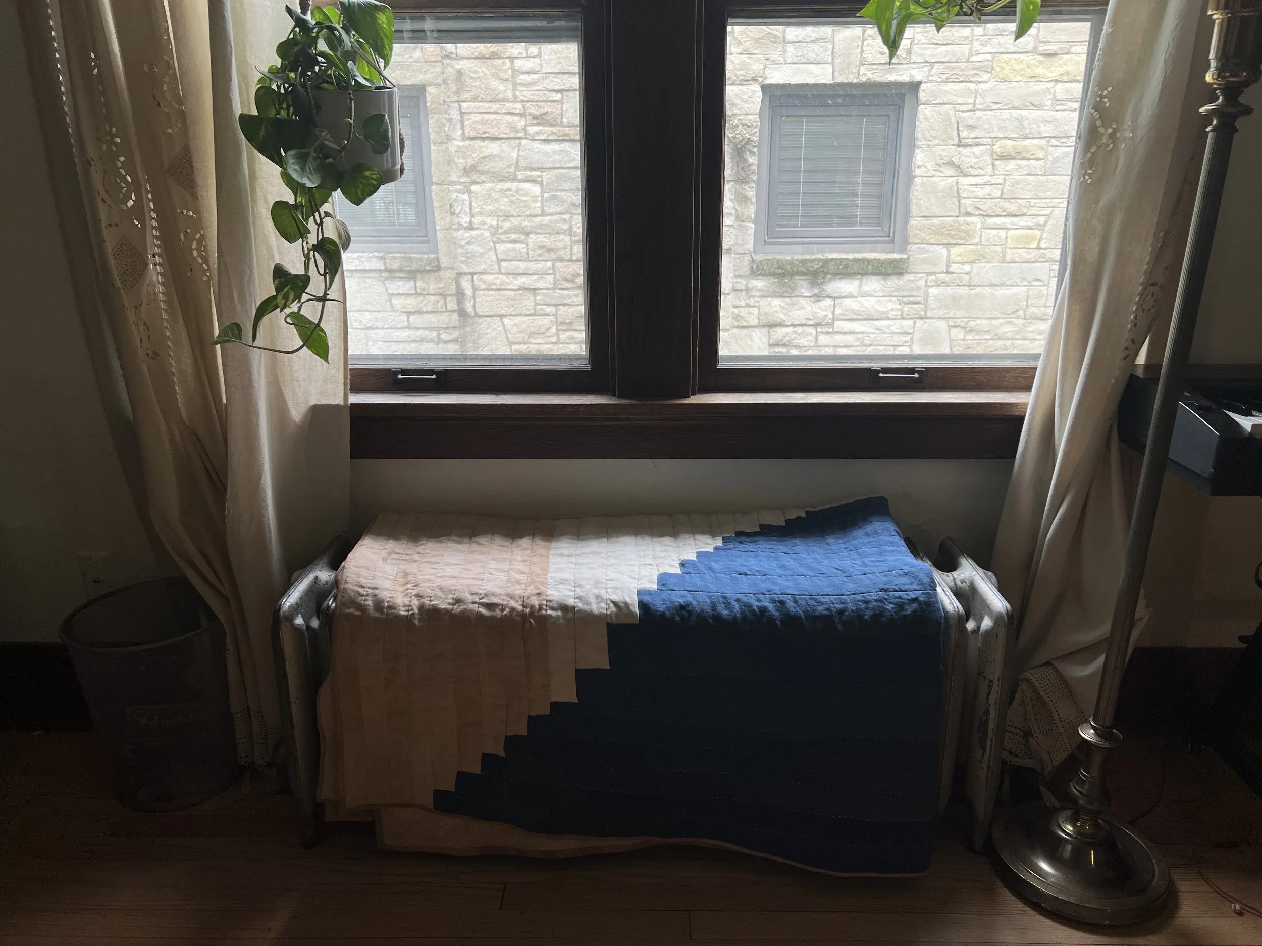 Indoor scene with a window partially covered by curtains, a potted plant hanging, a radiator with folded fabric on top, and a floor lamp.