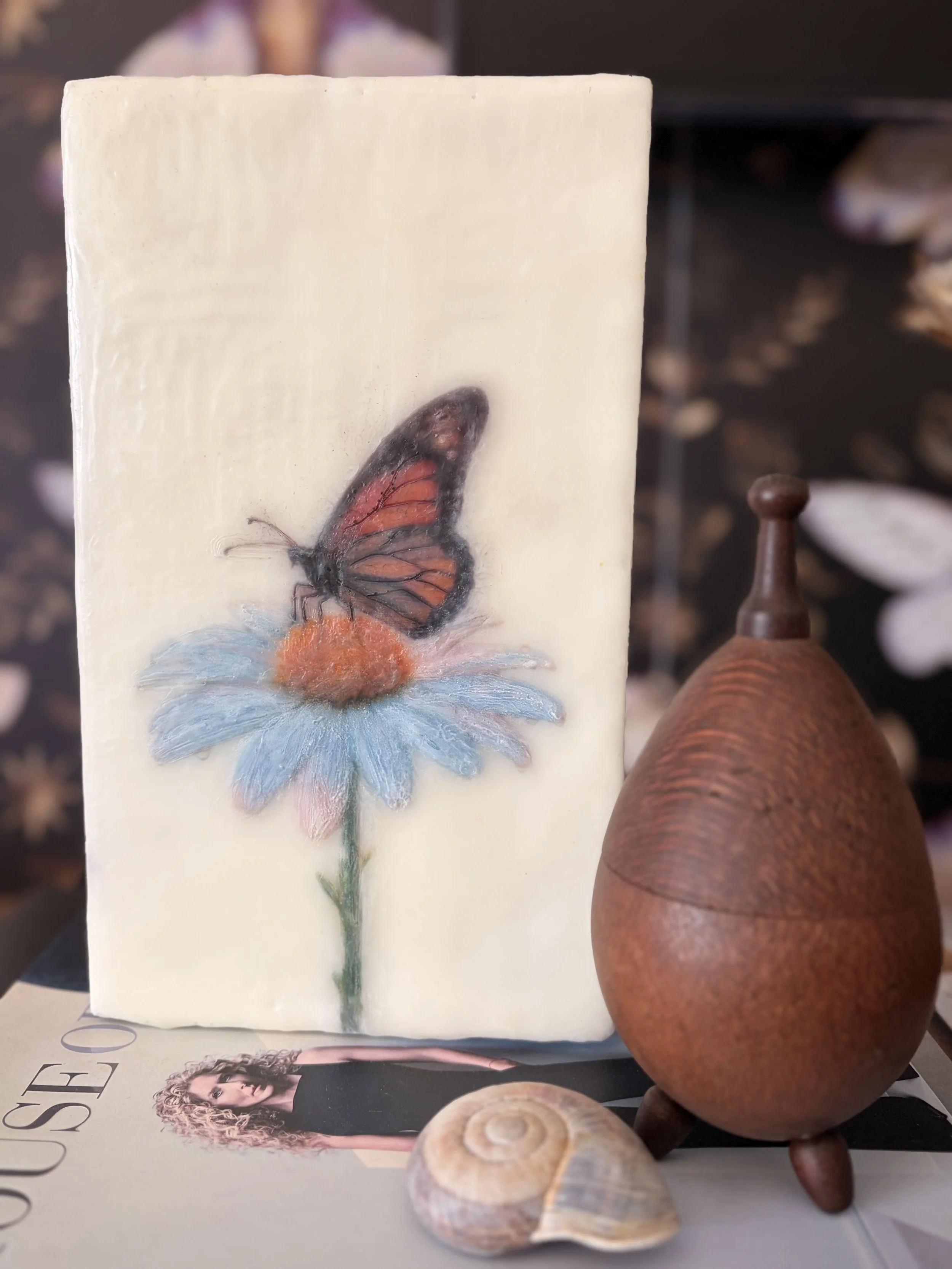 A warm orange butterfly perched on a pale blue daisy, created with encaustic wax and mixed media, featuring rich texture and soft botanical details.