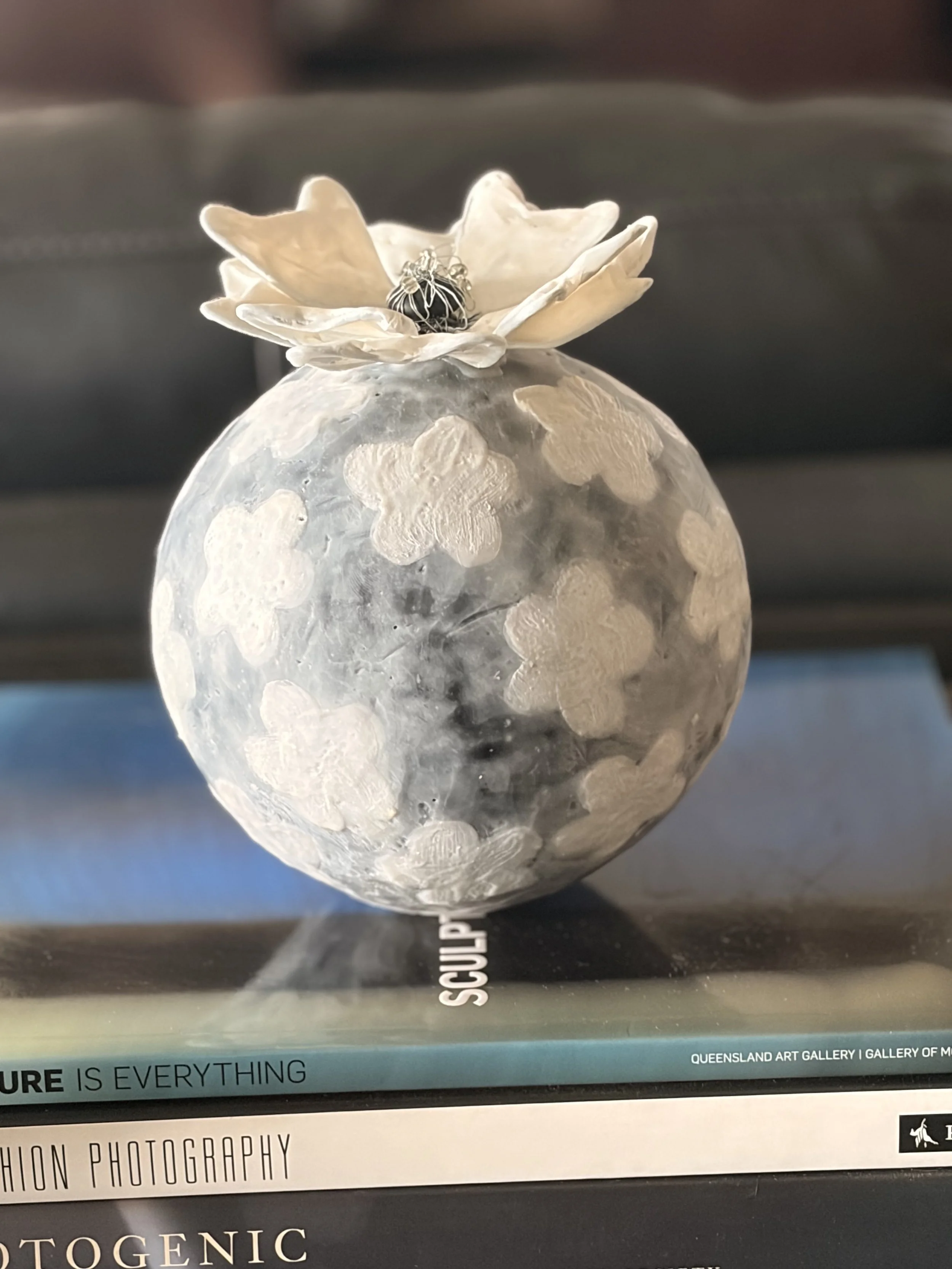 Encaustic wax and mixed media sculpture featuring a round grey textured form with raised white floral patterns and a sculptural white flower on top, displayed as a decorative art object.