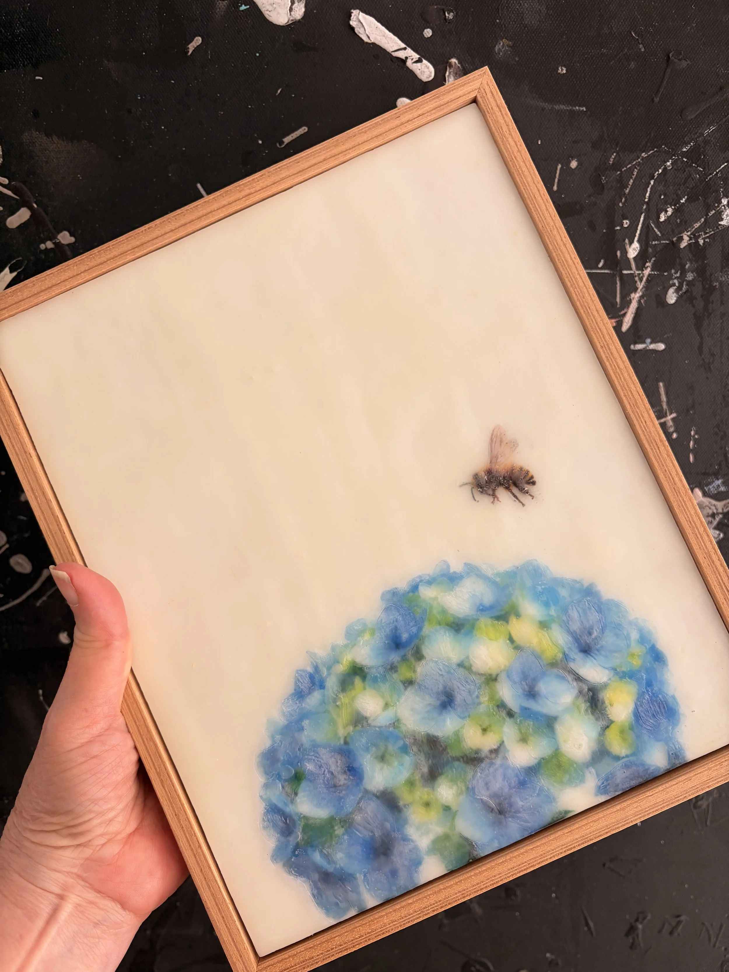 Original encaustic wax artwork featuring a honeybee hovering above blue hydrangea flowers, framed in Tasmanian oak with soft translucent beeswax layers.
