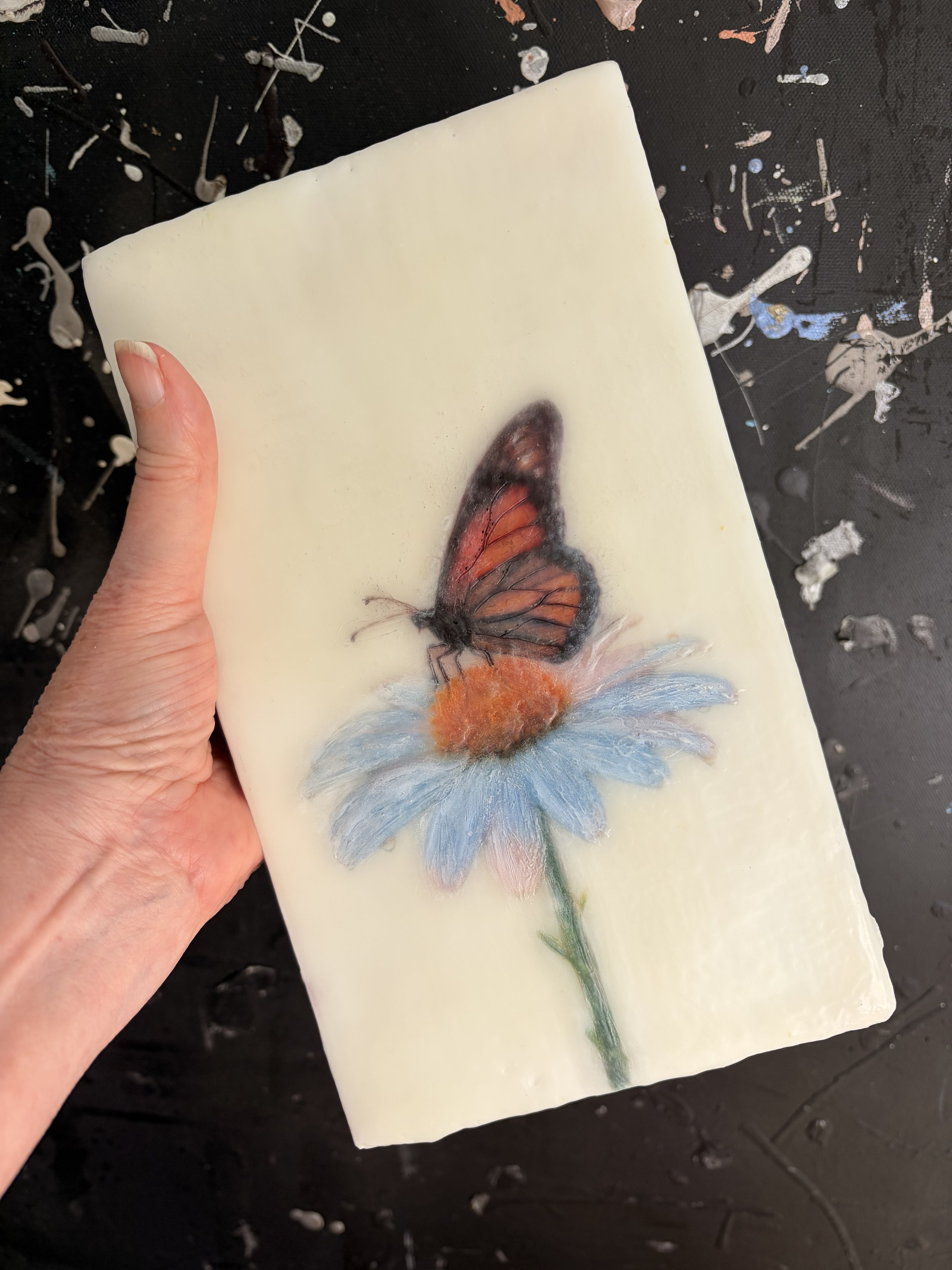 A warm orange butterfly perched on a pale blue daisy, created with encaustic wax and mixed media, featuring rich texture and soft botanical details.