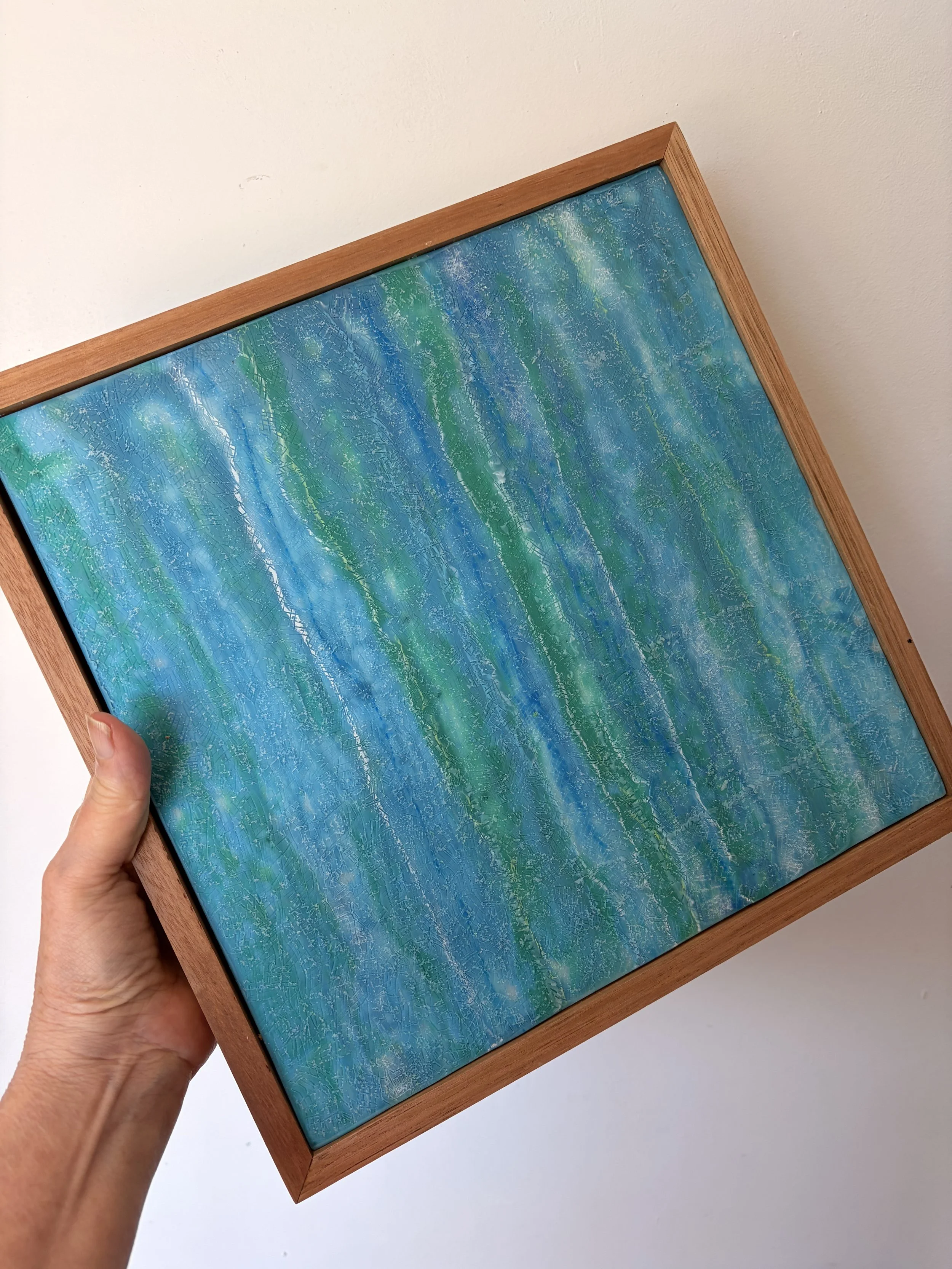 “A square 30 x 30cm encaustic wax and mixed media artwork featuring layered blue and green abstract textures, framed in natural timber.”