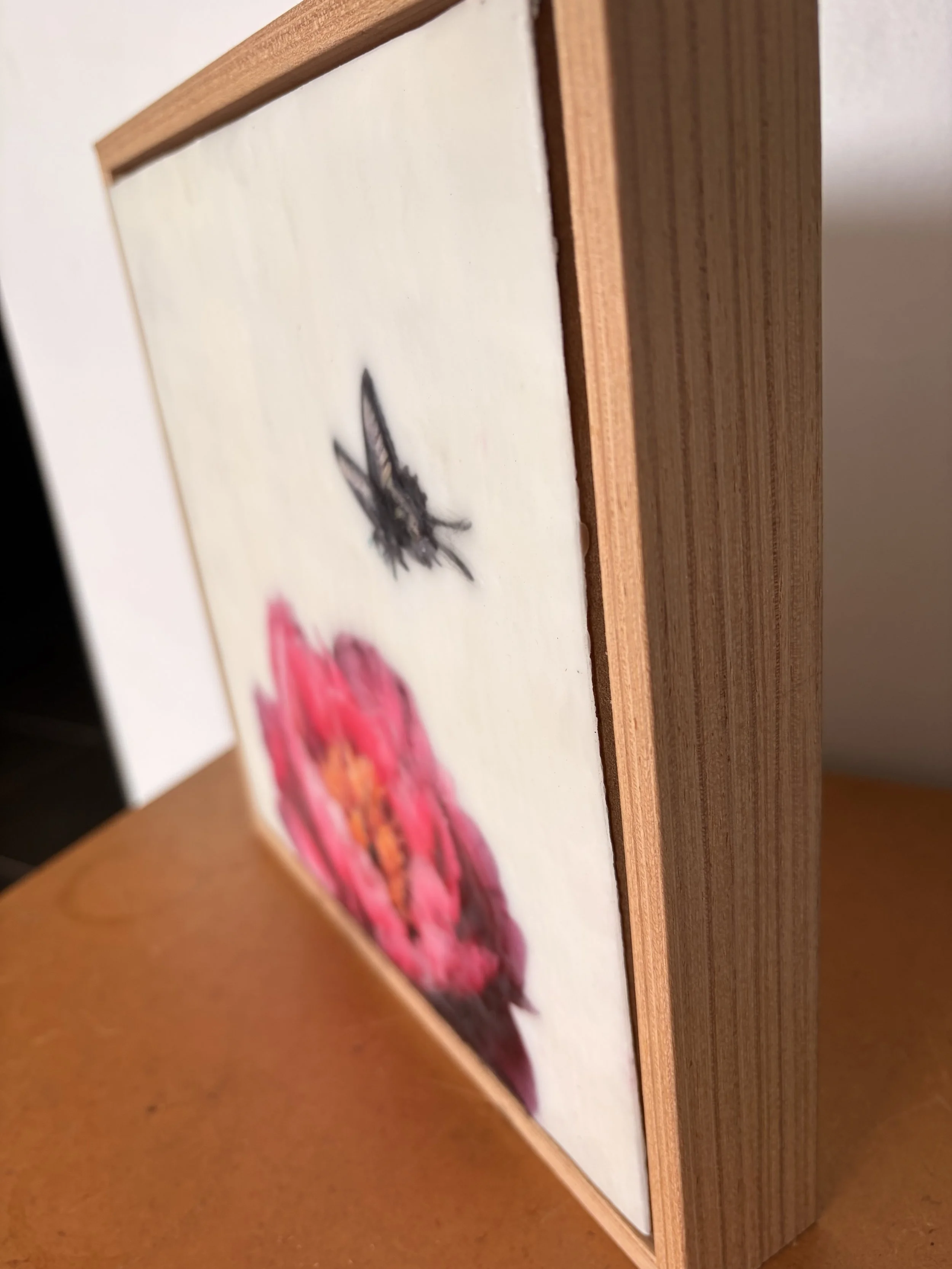 Encaustic wax artwork of a soft pink flower with a delicate butterfly hovering above, created with layered wax textures and gentle blended tones.