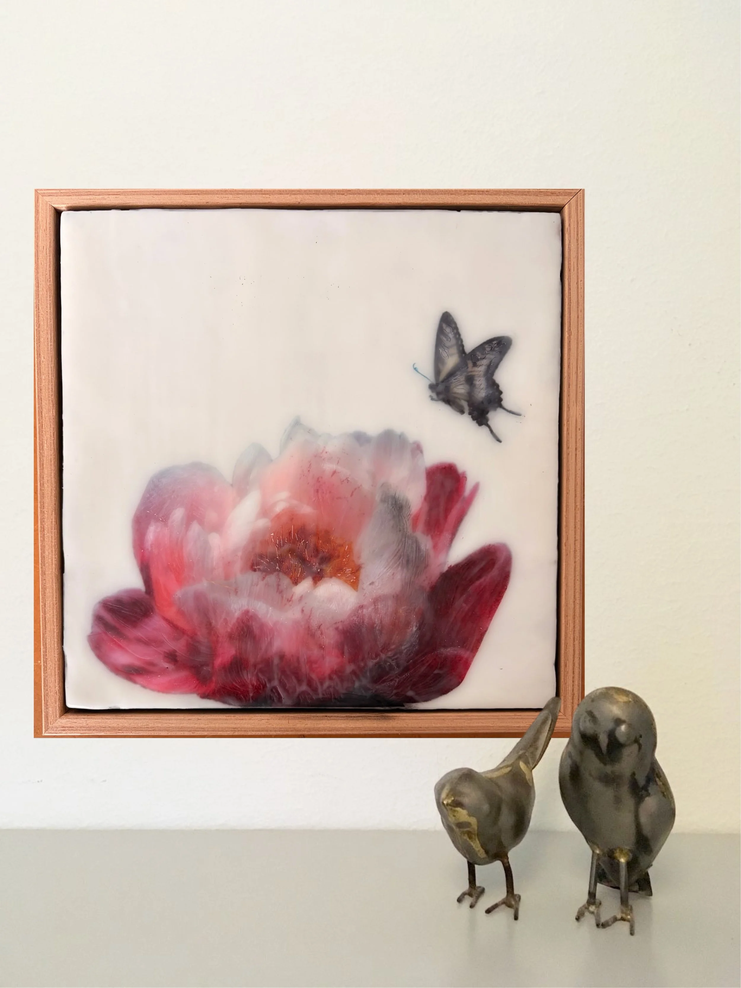 Encaustic wax artwork of a soft pink and crimson flower with a butterfly hovering above, featuring layered wax textures and a gentle, dreamlike finish.