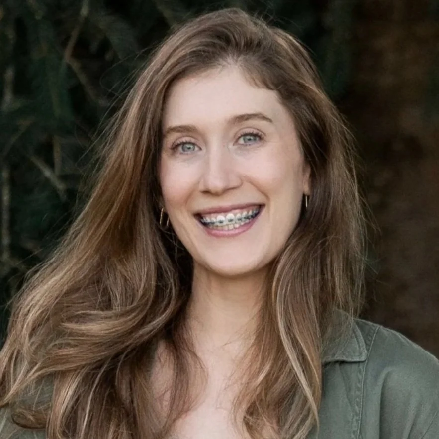 Portrait of a young woman with long, wavy brown hair, smiling, wearing braces and hoop earrings, outdoors with blurred greenery in the background.