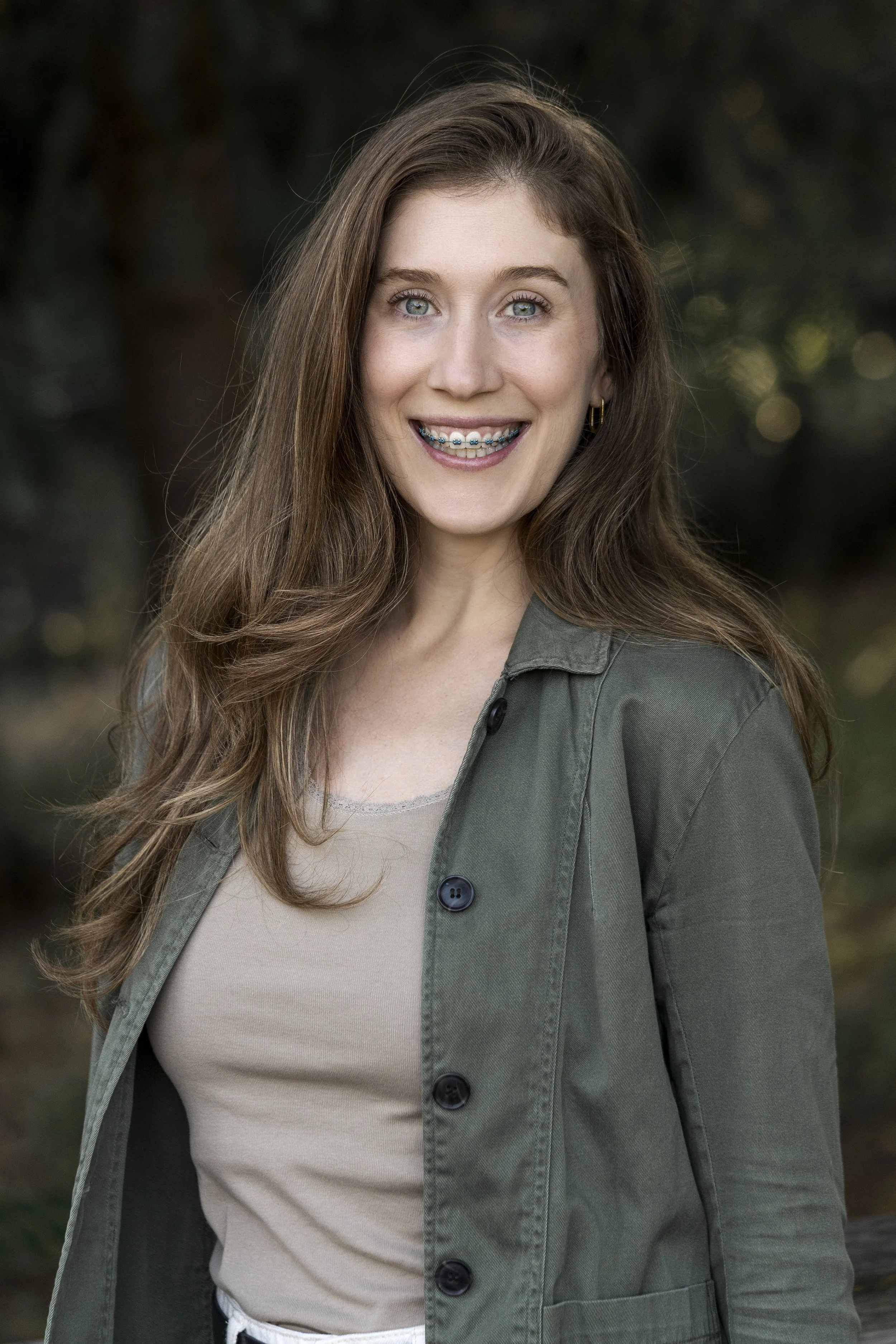 A woman with long brown hair and blue eyes smiling outdoors, wearing a beige top and a green jacket.