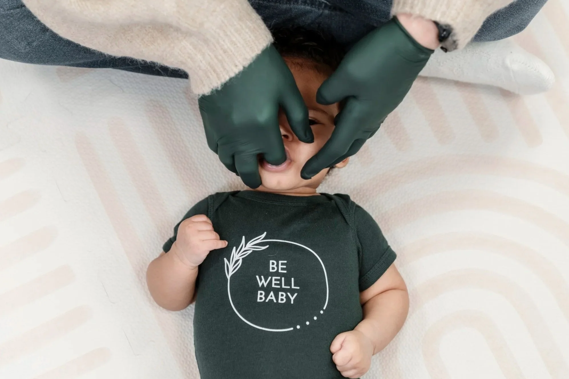 A person wearing green gloves checks a baby's mouth while the baby lies on a soft blanket, wearing a dark green onesie with the words 'BE WELL BABY' and a decorative circle with a leaf.