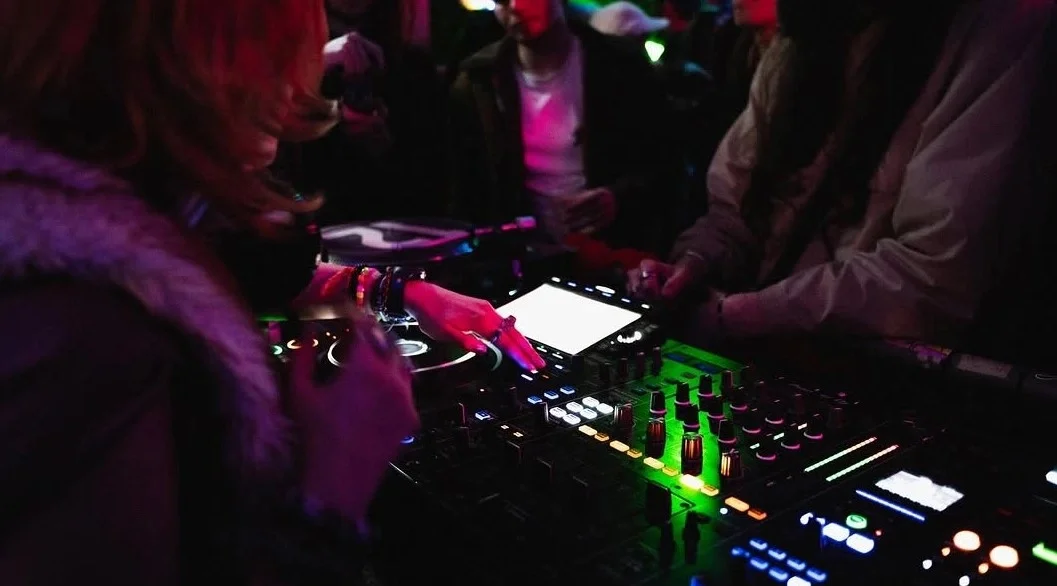 A DJ is mixing music at the Oculist Brighton with colorful lights, surrounded by people.