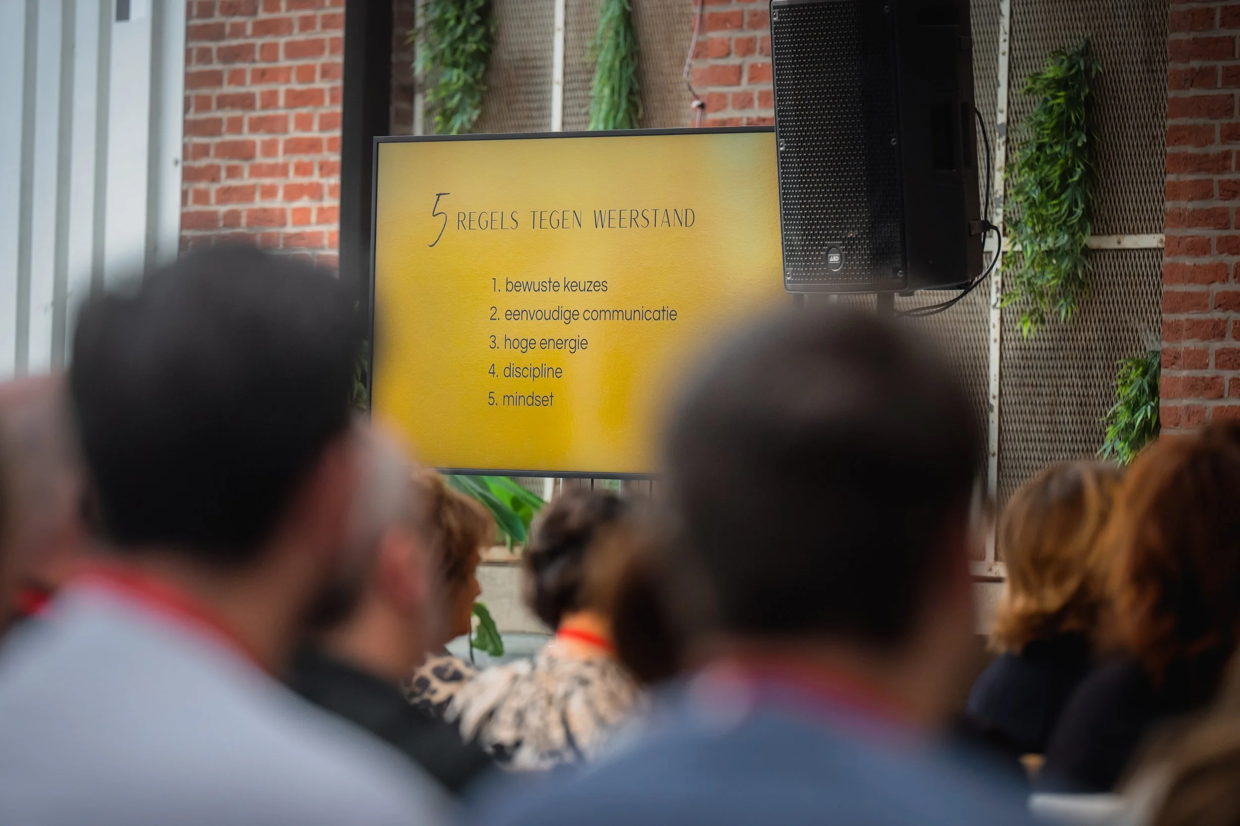 Presentatie met titel '5 regels tegen weerstand' op een scherm, lijst met vijf punten: 1. bewuste keuzes, 2. eenvoudige communicatie, 3. hoge energie, 4. discipline, 5. mindset; publiek zit in een zaal met planten en een bakstenen muur op de achtergrond.